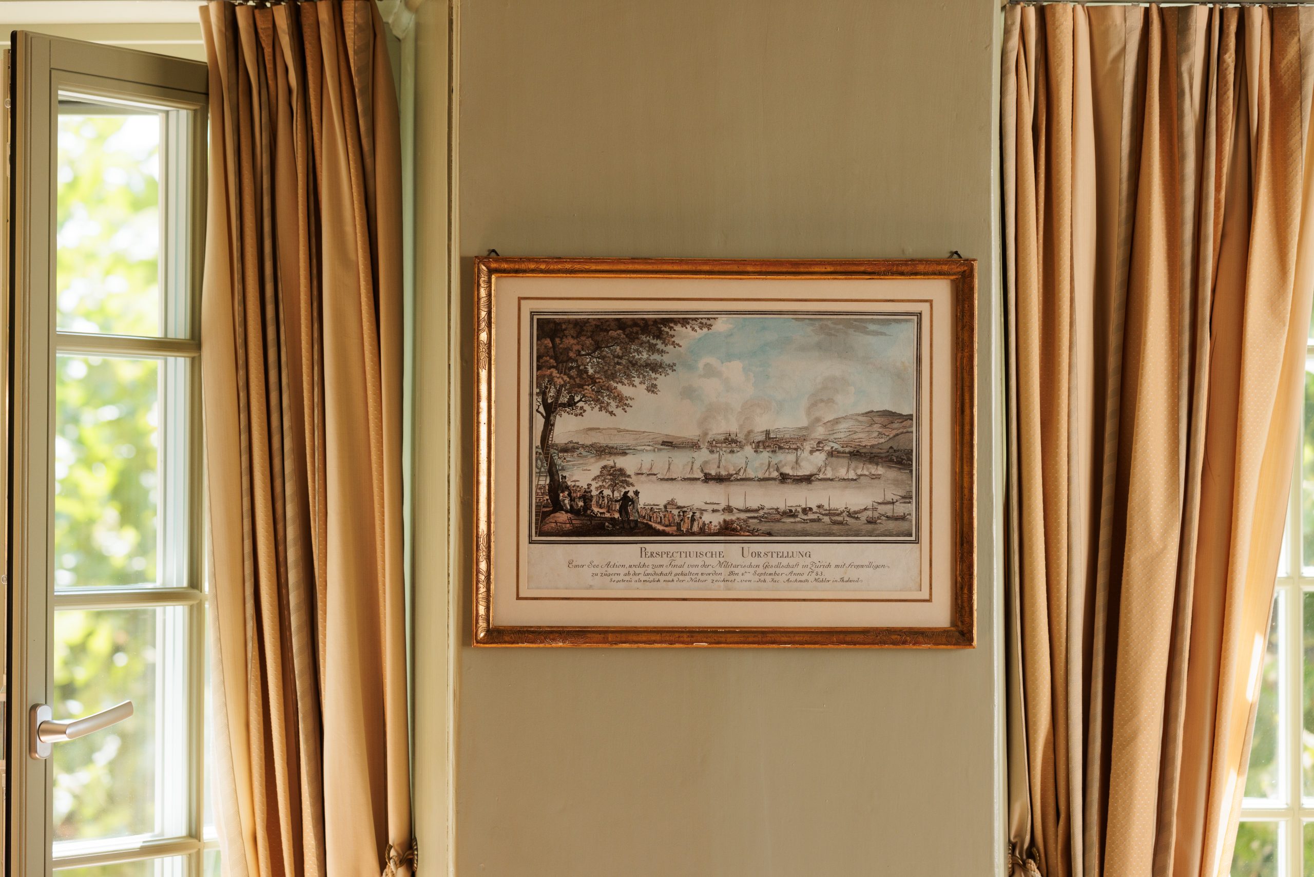 Ein gerahmtes Vintage-Gemälde mit Schiffen im Hafen hängt an einer beigen Wand zwischen zwei Fenstern mit hellbraunen Vorhängen im Hotel Sonne. Das Sonnenlicht beleuchtet sanft diese von der Kunst inspirierte Szene.