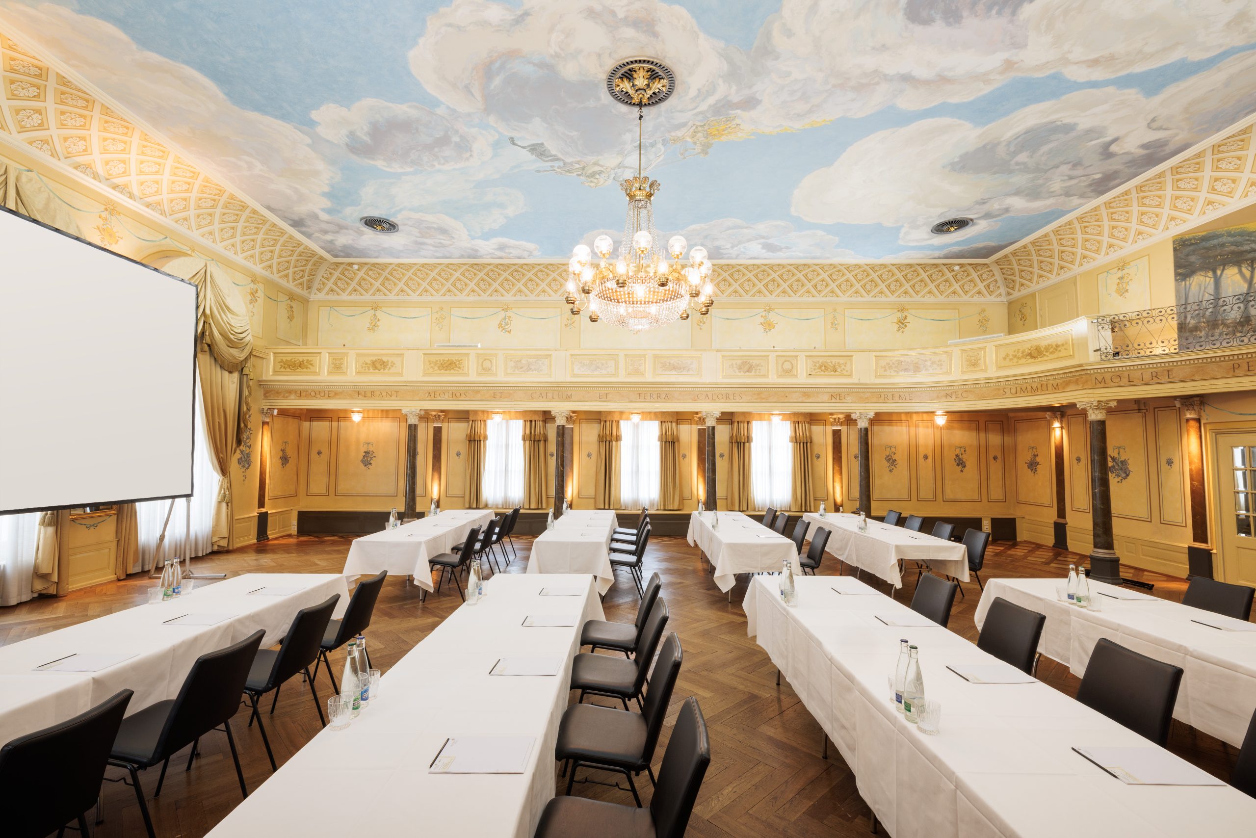 Eleganter Konferenzraum im Hotel Sonne mit verschnörkelten gelben Wänden, großen Fenstern, einem Kronleuchter und einer bemalten Himmelsdecke - perfekt für Ihr nächstes Seminar. Die Tische in U-Form sind mit Notizblöcken und Wasserflaschen für jeden Teilnehmer ausgestattet.