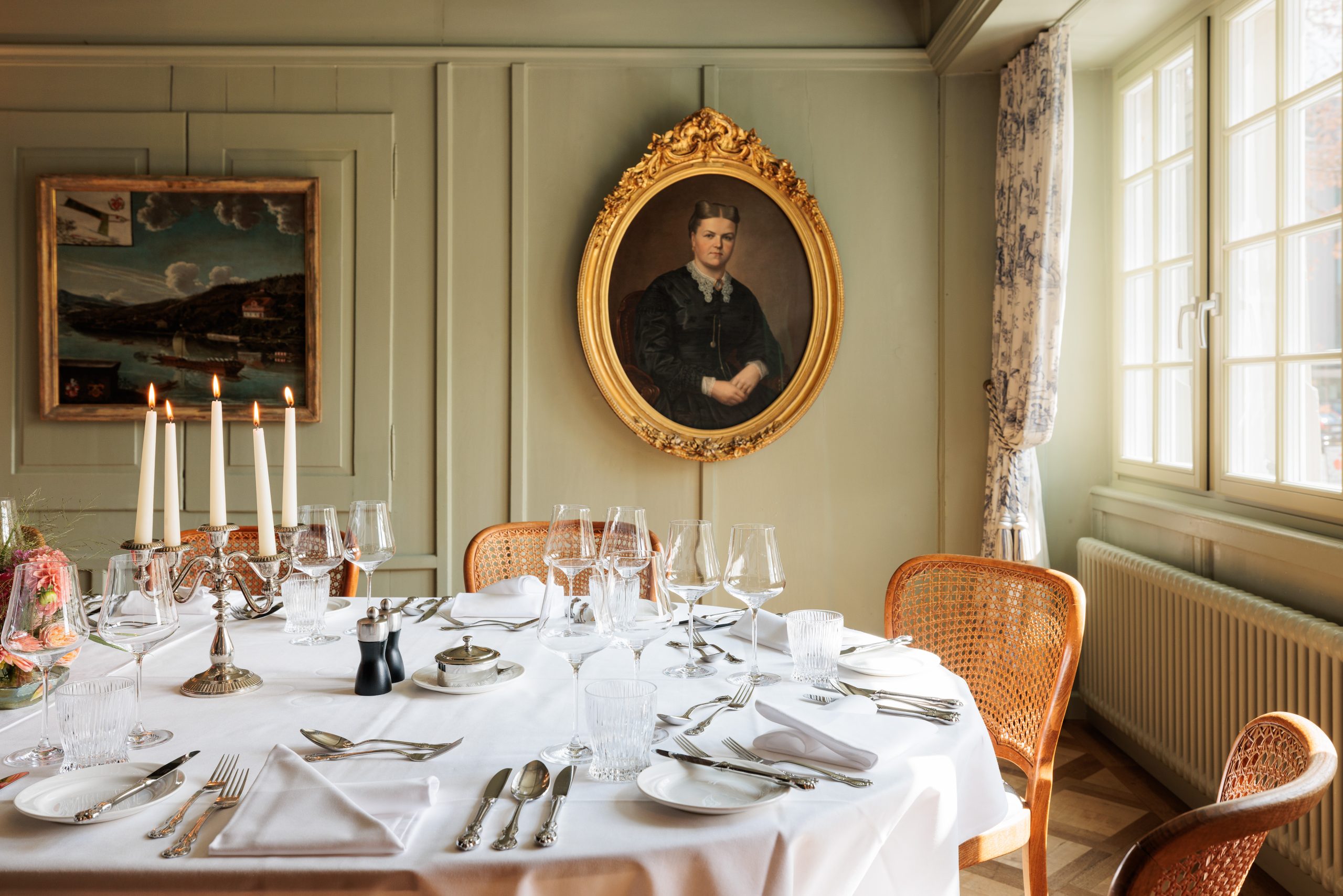 Elegantes Esszimmer im Event Hotel Sonne mit einem runden Tisch, der für eine Mahlzeit gedeckt ist, mit weißer Tischdecke, Kerzen, Gläsern und Besteck. Goldgerahmte Porträt- und Landschaftsgemälde schmücken die Wand neben den großen Fenstern.