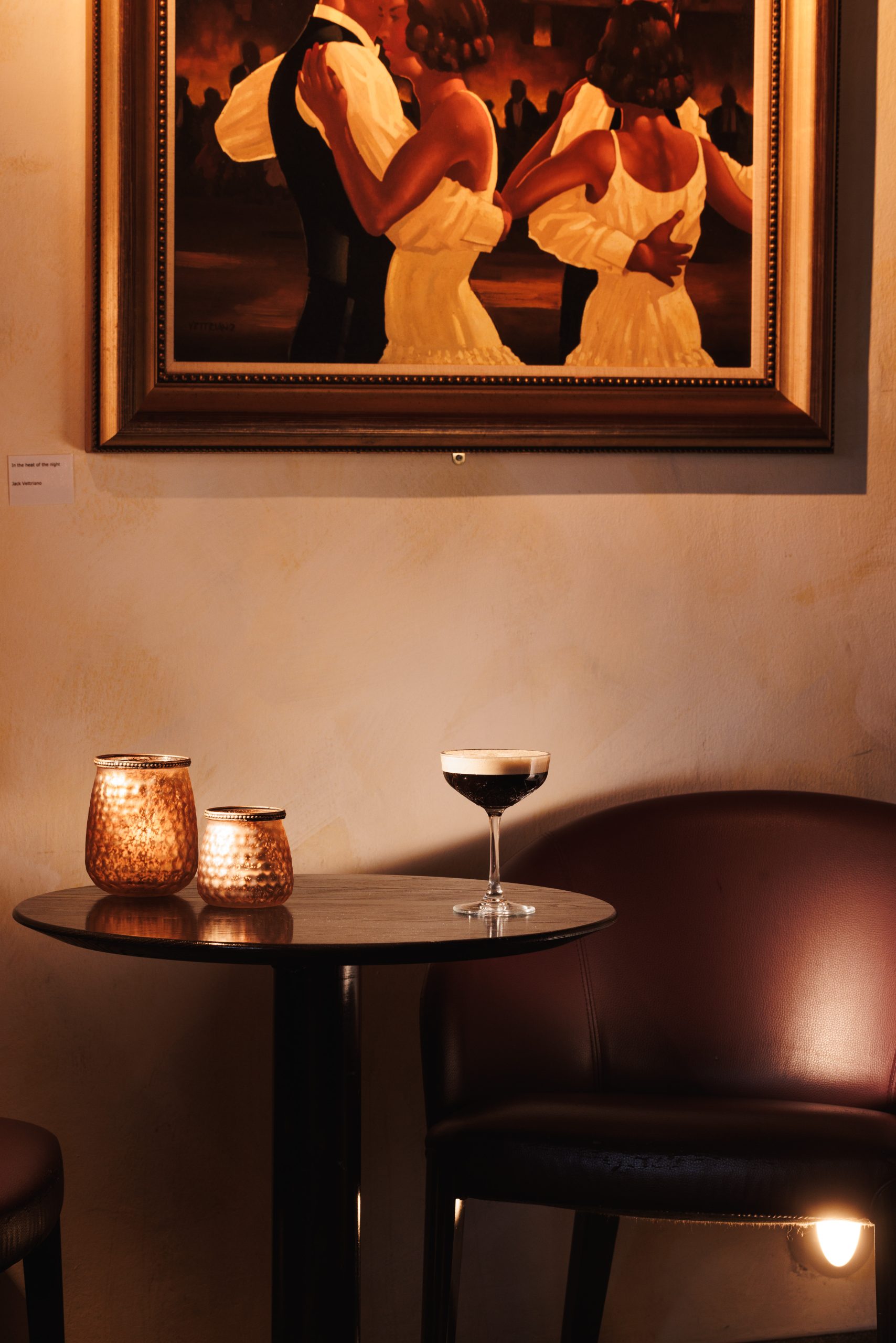 Ein kleiner Cocktailbar-Tisch mit zwei brennenden Kerzen und einem Cocktailglas steht zwischen zwei braunen Lederstühlen. Darüber zeigt ein gerahmtes Bild Paare in eleganter Kleidung, die eng beieinander tanzen und ein warmes und intimes Hotelambiente schaffen.