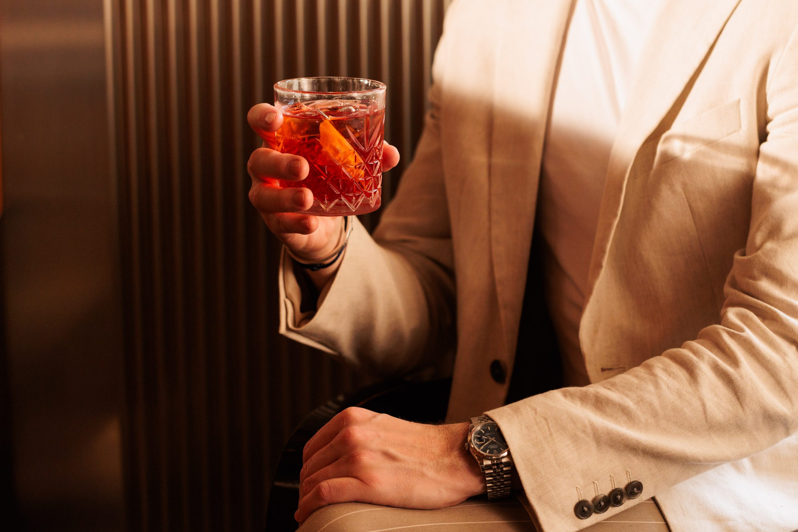 Eine Person in beigefarbenem Anzug und weißem Hemd sitzt mit einem Turmbar-Cocktail in der Hand, der mit einer Orangenscheibe garniert ist. Nur der Oberkörper und die Hände sind zu sehen, wobei eine Armbanduhr hervorsticht, die gegen eine gerippte Wand gelehnt ist.