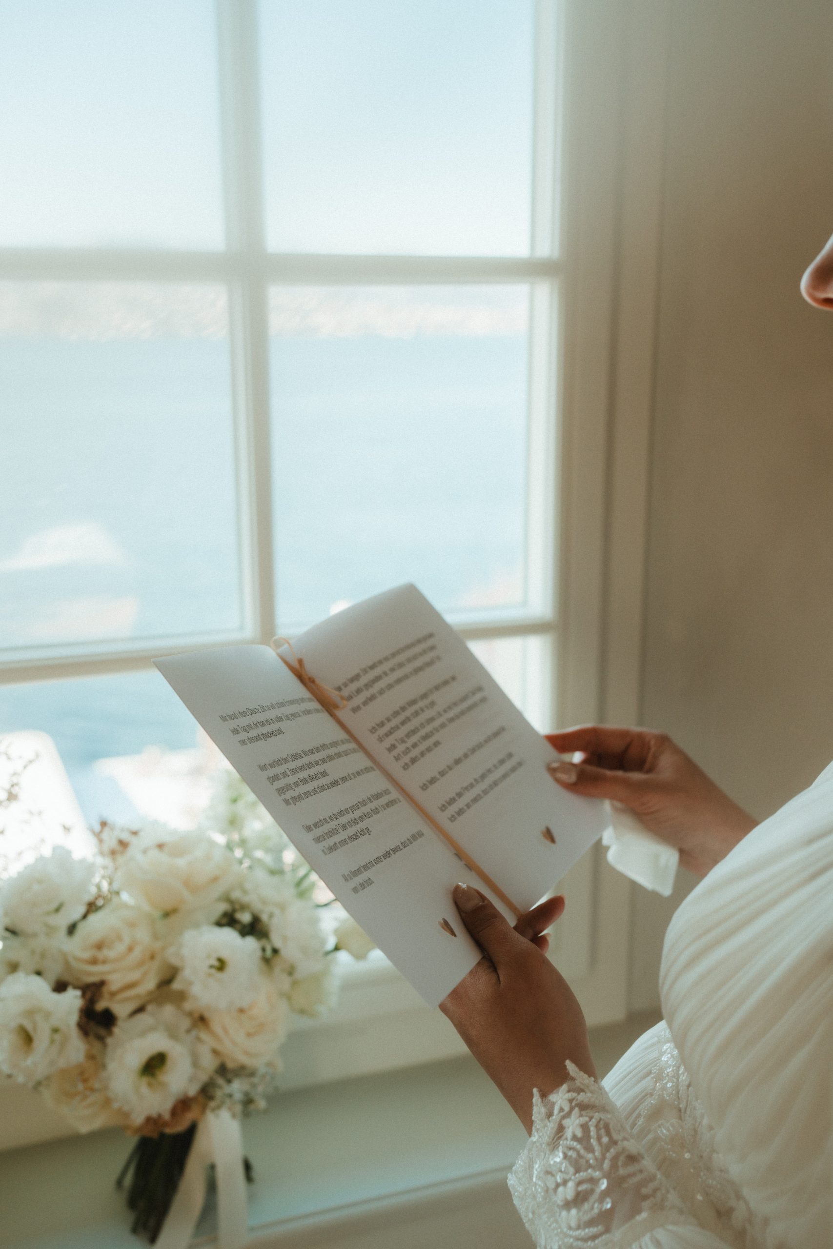 Eine Braut in einem weißen Spitzenkleid liest ein Büchlein am Fenster ihrer Hochzeit am See; draußen bietet sich ein herrlicher Blick auf das Wasser, und auf der Fensterbank daneben liegt ein Strauß weißer Blumen.