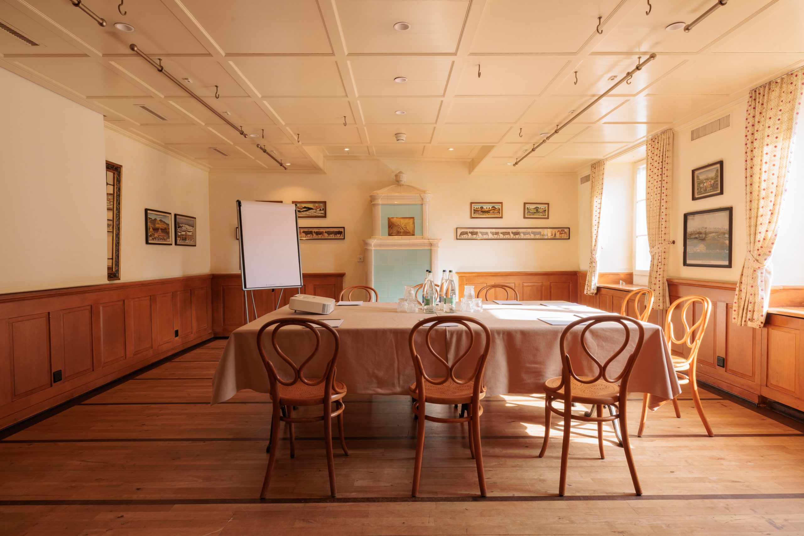 Ein sonnendurchfluteter Seminarraum im Hotel Sonne ist mit einem mit beigem Tuch bedeckten Tisch, sechs Holzstühlen, Glasflaschen, Gläsern, einem Flipchart und gerahmten Kunstwerken an holzgetäfelten Wänden ausgestattet. Große Fenster lassen viel natürliches Licht herein.