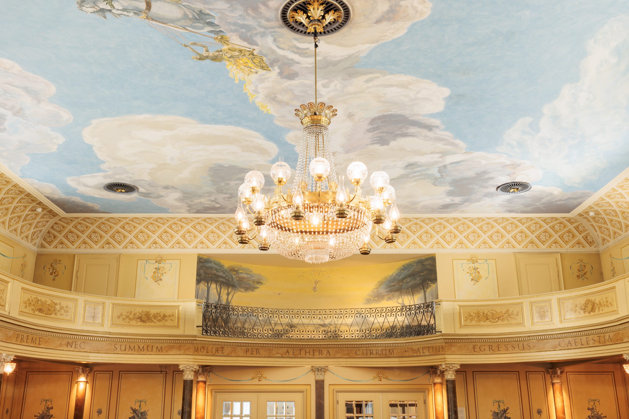 Im Festsaal des Hotels Sonne hängt ein prächtiger Kronleuchter von einer mit Wolken und Himmel bemalten Decke über einem kunstvoll gestalteten Raum mit gelben Wänden, klassischen Wandmalereien und dekorativen Stuckarbeiten - perfekt für Ihre nächste Veranstaltung.