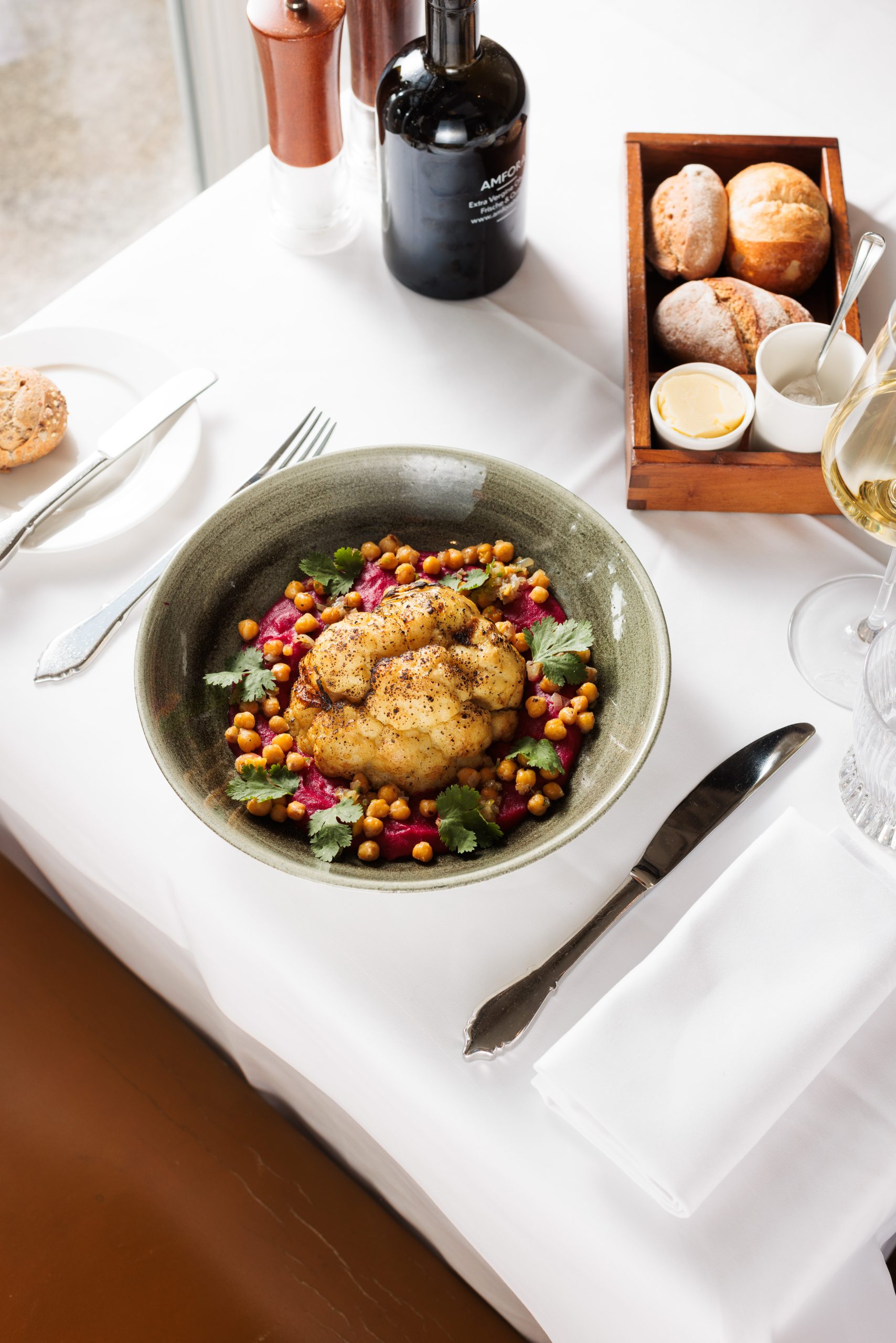 Eine Schale mit gebratenem Hähnchen auf einem Bett aus Kichererbsen und Gemüse steht auf einem weißen Tischtuch in der Sonnengalerie, umgeben von Brötchen, Butter, einem Weinglas, einer Flasche und Besteck, bereit für ein köstliches Mahl.