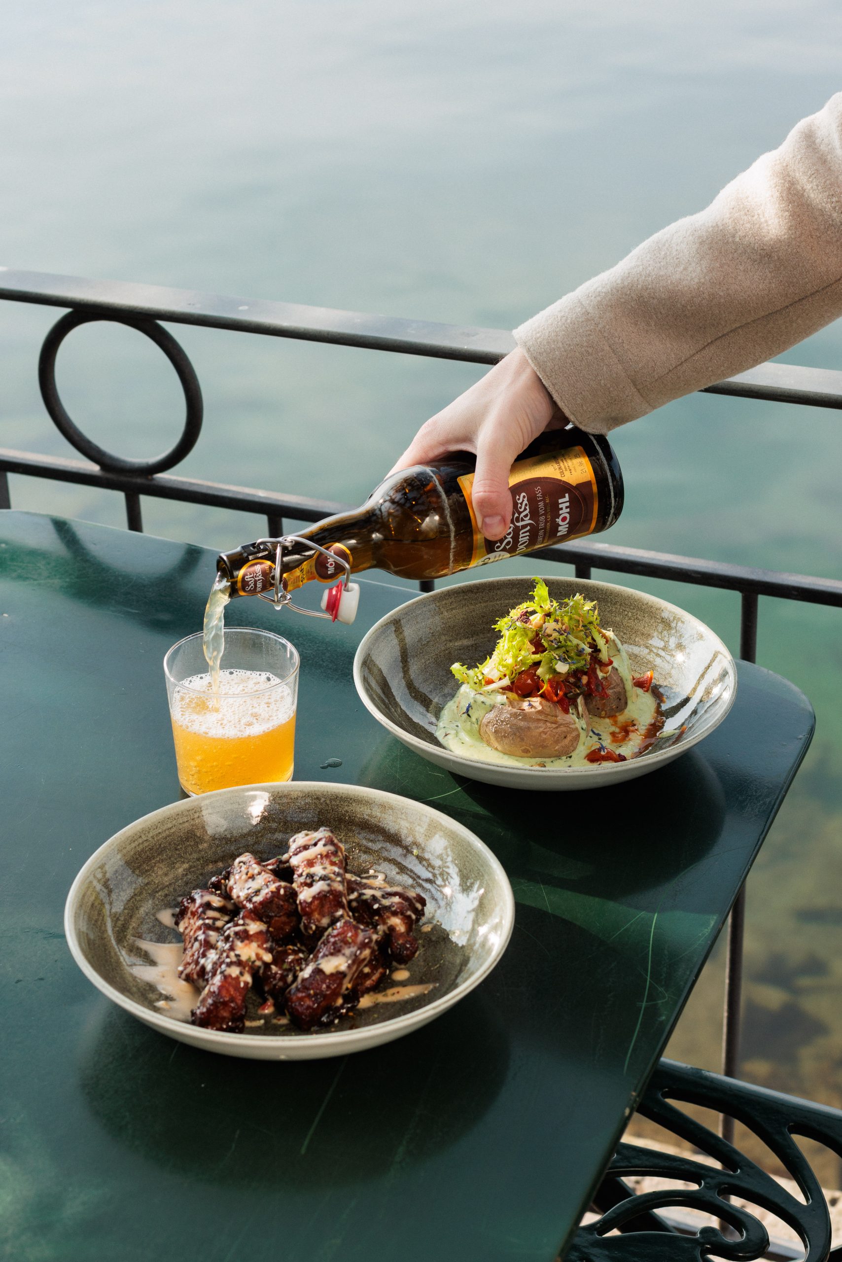 Eine Person gießt Bier in ein Glas an einem grünen Tisch im Biergarten-Stil, der mit saftigen Hähnchenflügeln und einem Salatteller gedeckt ist, alles in der Nähe eines malerischen Geländers am Wasser.