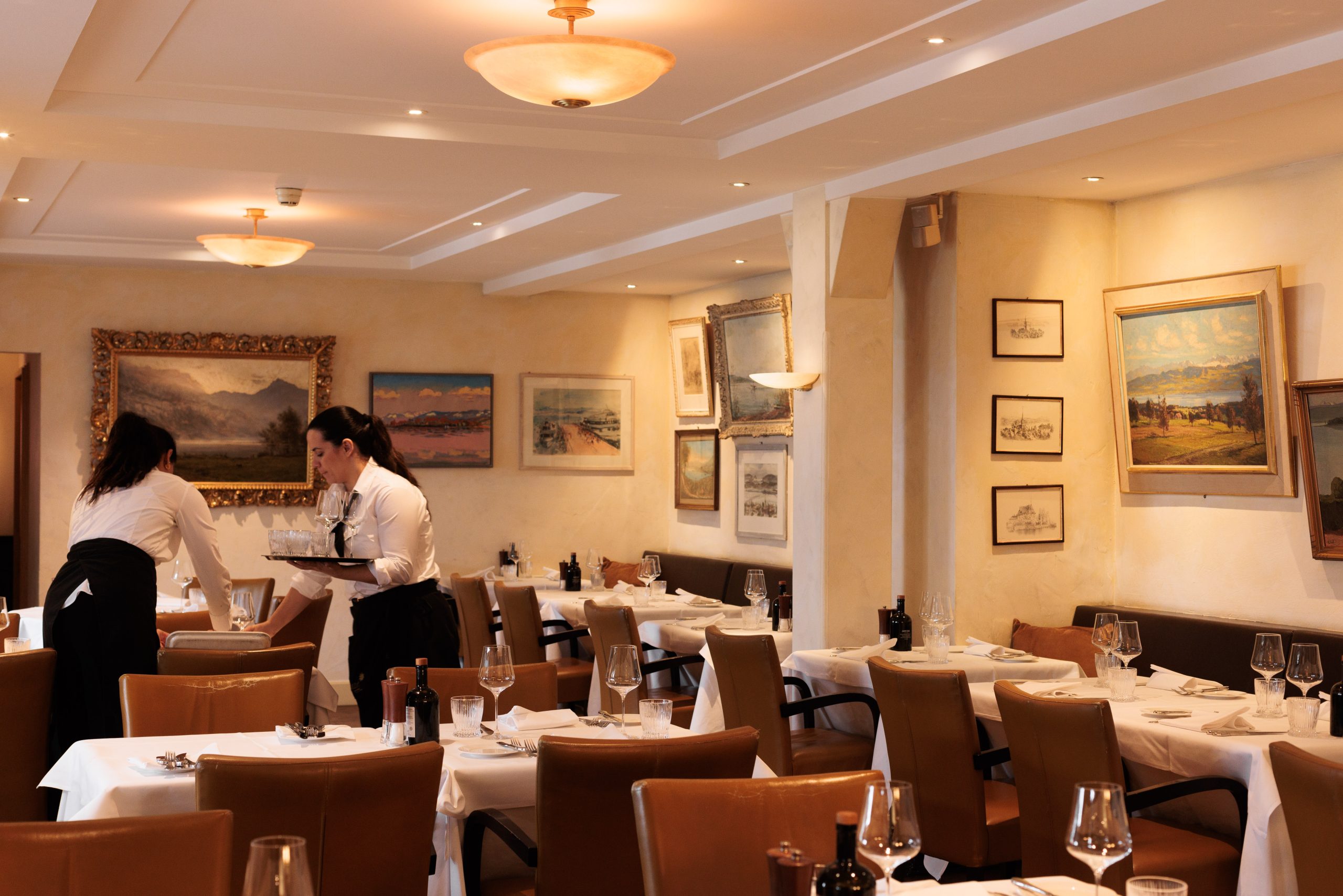 Zwei Kellner in weißen Hemden und schwarzen Schürzen decken die Tische im eleganten Restaurant Sonnengalerie mit weißen Tischtüchern, braunen Stühlen und gerahmten Gemälden an den Wänden bei warmer Beleuchtung.
