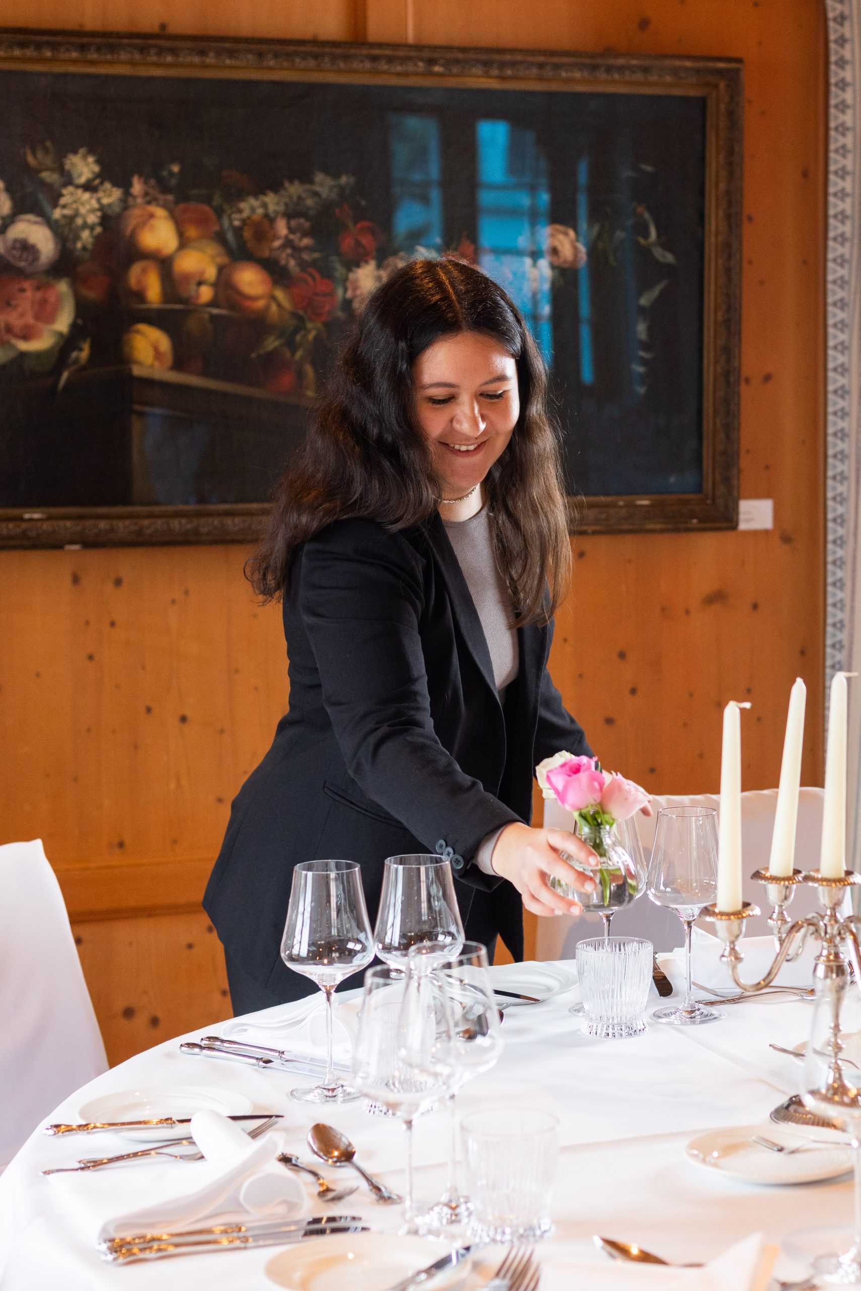 Eine Frau mit langen dunklen Haaren und schwarzem Blazer stellt eine kleine Vase mit rosa Rosen auf einen formell gedeckten Esstisch im Hotel Sonne, umgeben von Kerzen und Gläsern in einem eleganten Raum mit holzgetäfelten Wänden und einem Obstgemälde.