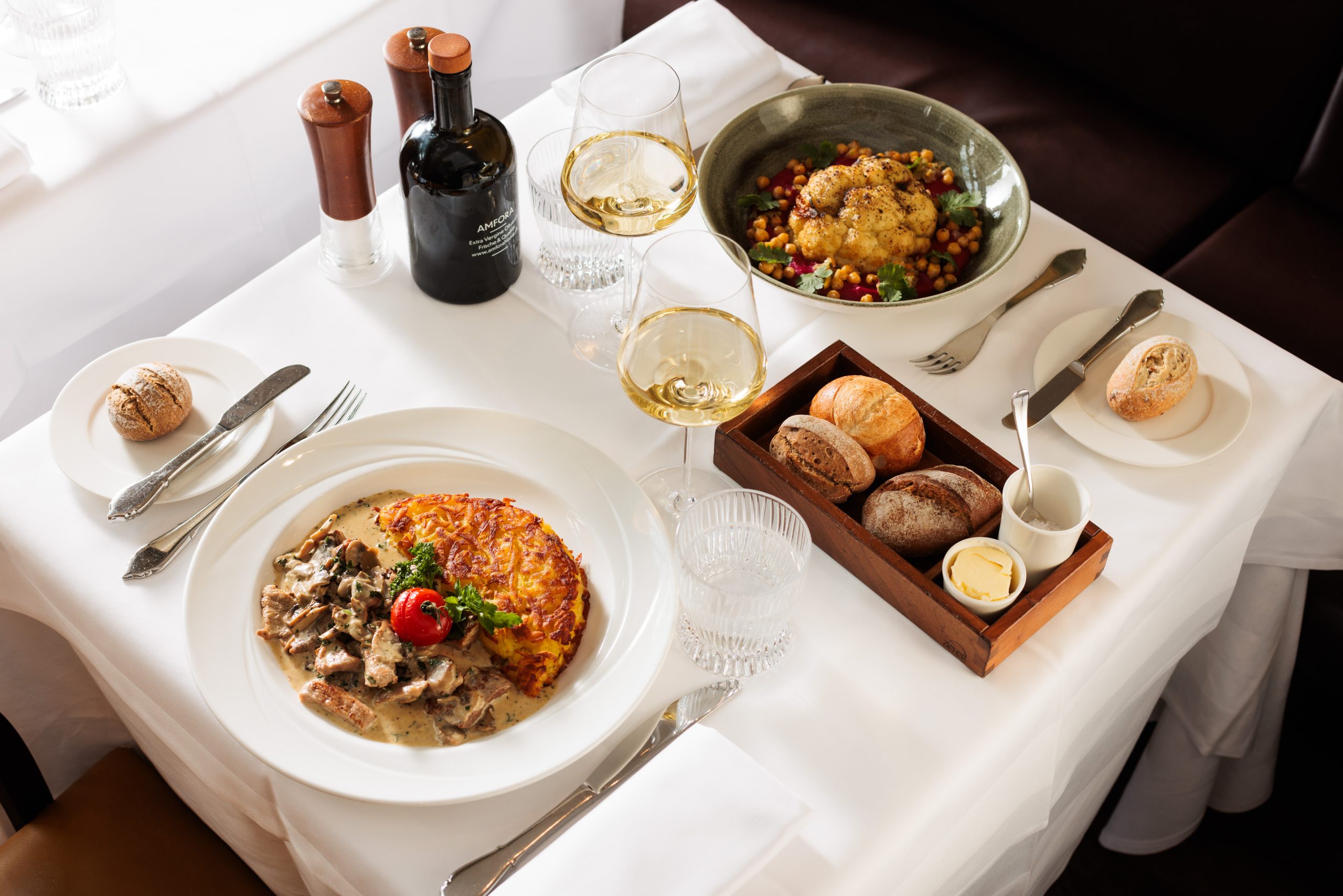 Ein gedeckter Tisch in der Sonnengalerie für zwei Personen mit Weingläsern, Brötchen, Butter, einem Teller Fleisch mit Soße und Rösti, einer Schüssel Salat mit Kichererbsen und Gewürzen auf einem weißen Tischtuch.