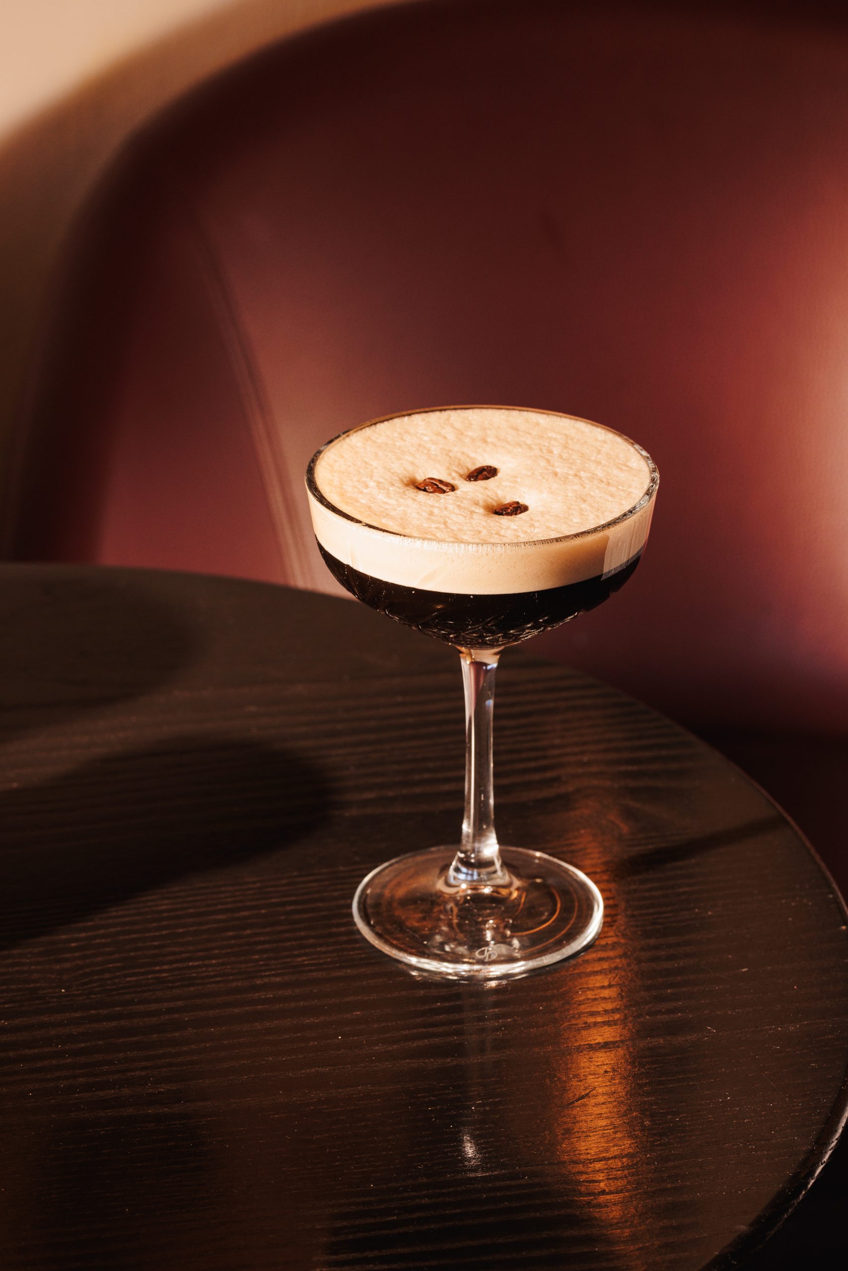 Ein Glas Espresso Martini Hotel Sonne mit Schaum und drei Kaffeebohnen steht elegant auf einem dunklen Holztisch vor einem kastanienbraunen Stuhl.