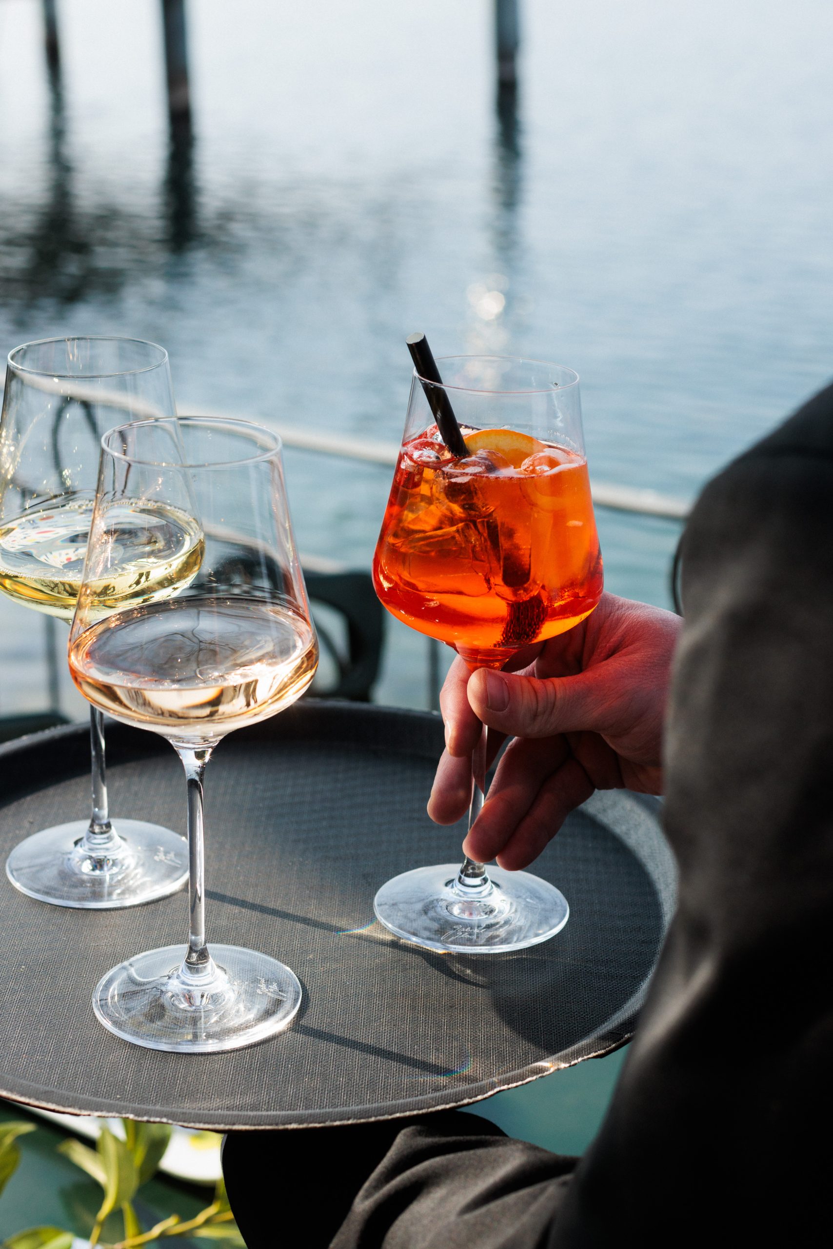 Ein Kellner in einem Biergarten hält ein Tablett mit drei Gläsern: eines mit einem Orangencocktail und einem Strohhalm und zwei mit Weiß- und Roséwein. Im Hintergrund sind Wasser und Holzpfähle zu sehen.