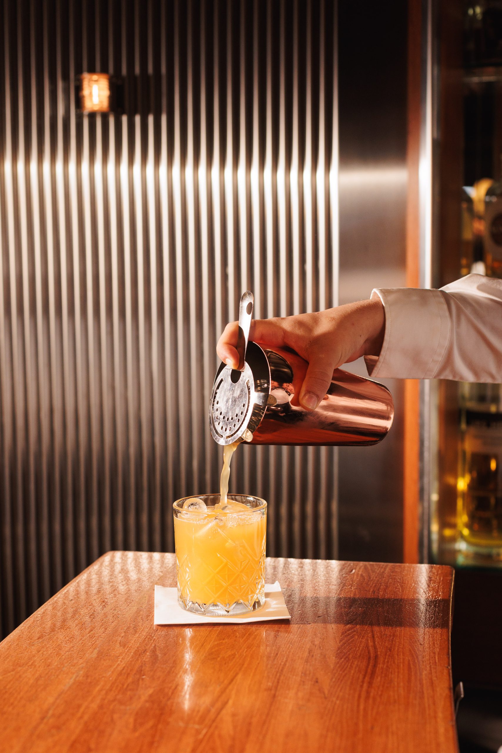 Ein Barkeeper der Turmbar gießt einen gelben Cocktail aus einem Shaker durch ein Sieb in ein Glas auf einer Serviette, das auf einer hölzernen Bar mit einer Wellblechwand im Hintergrund steht.
