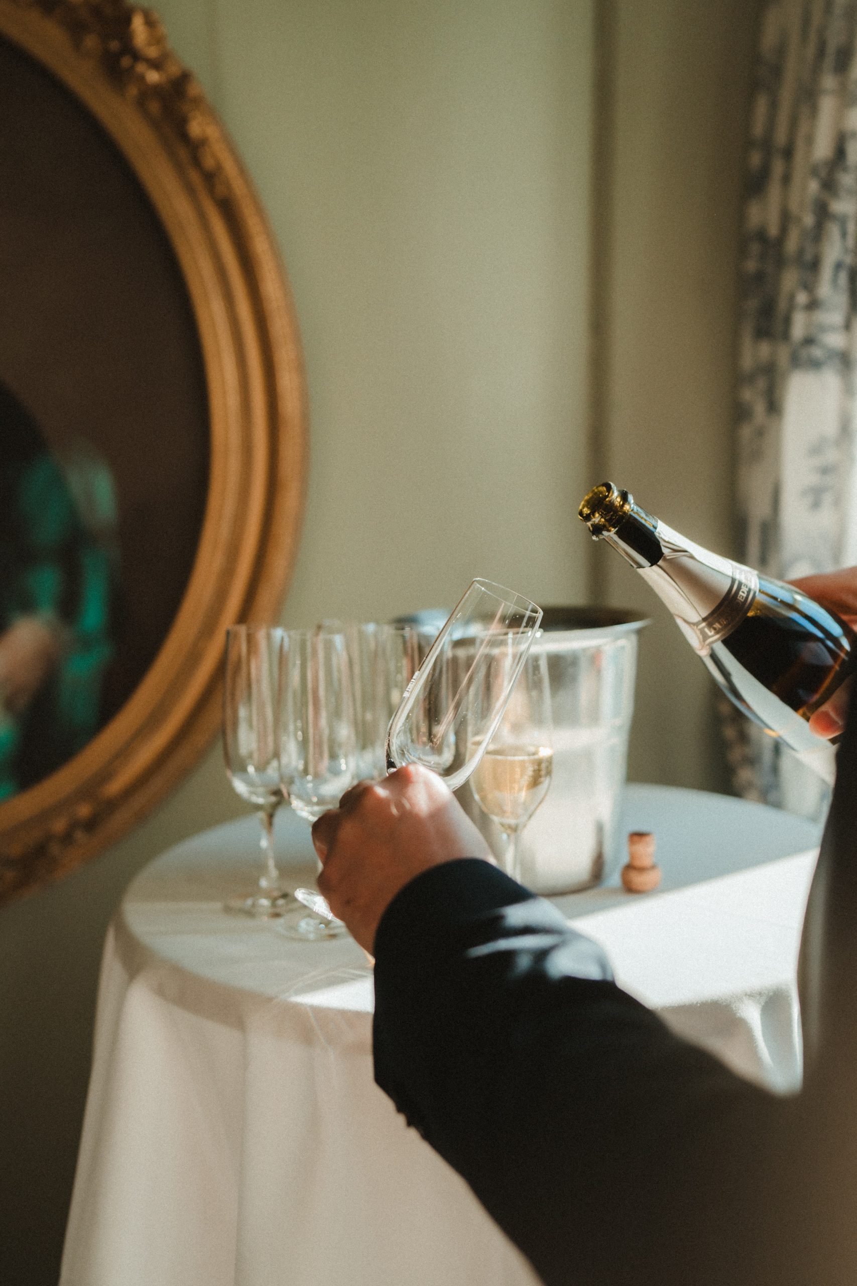 Eine formell gekleidete Person schenkt Champagner in ein Glas neben leeren Gläsern und einem Eiskübel auf einem runden Tisch mit weißem Tischtuch ein - eine elegante Szene, wie geschaffen für eine Hochzeit am Zürichsee. Ein goldgerahmtes Gemälde ziert die Wand daneben.