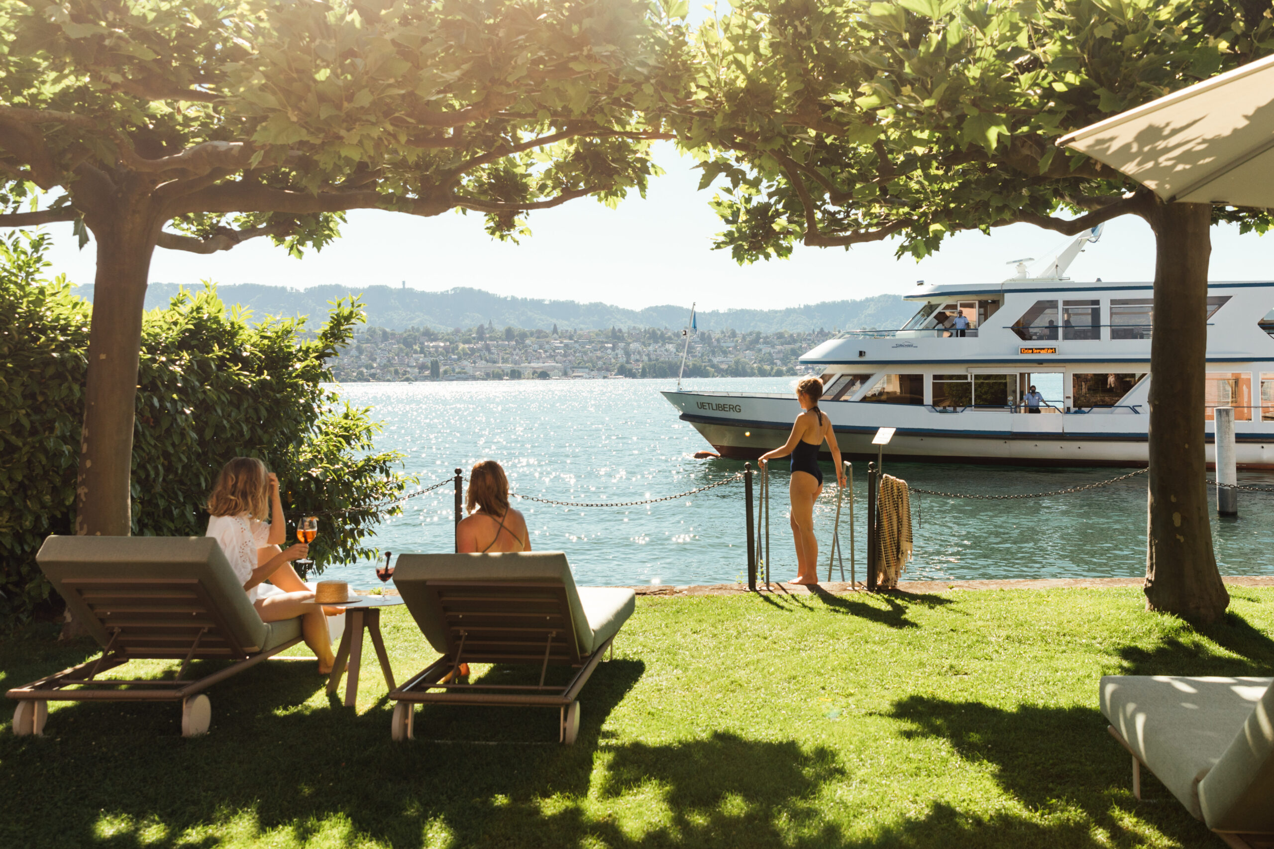 Auf der Seewiese Küsnacht entspannen sich zwei Personen auf Liegestühlen unter Bäumen am See, in der Hand Getränke. Eine weitere Person in Badebekleidung steht am Wasser und beobachtet ein grosses weisses Boot, das am Hotel Sonne und den fernen Hügeln über den See gleitet.