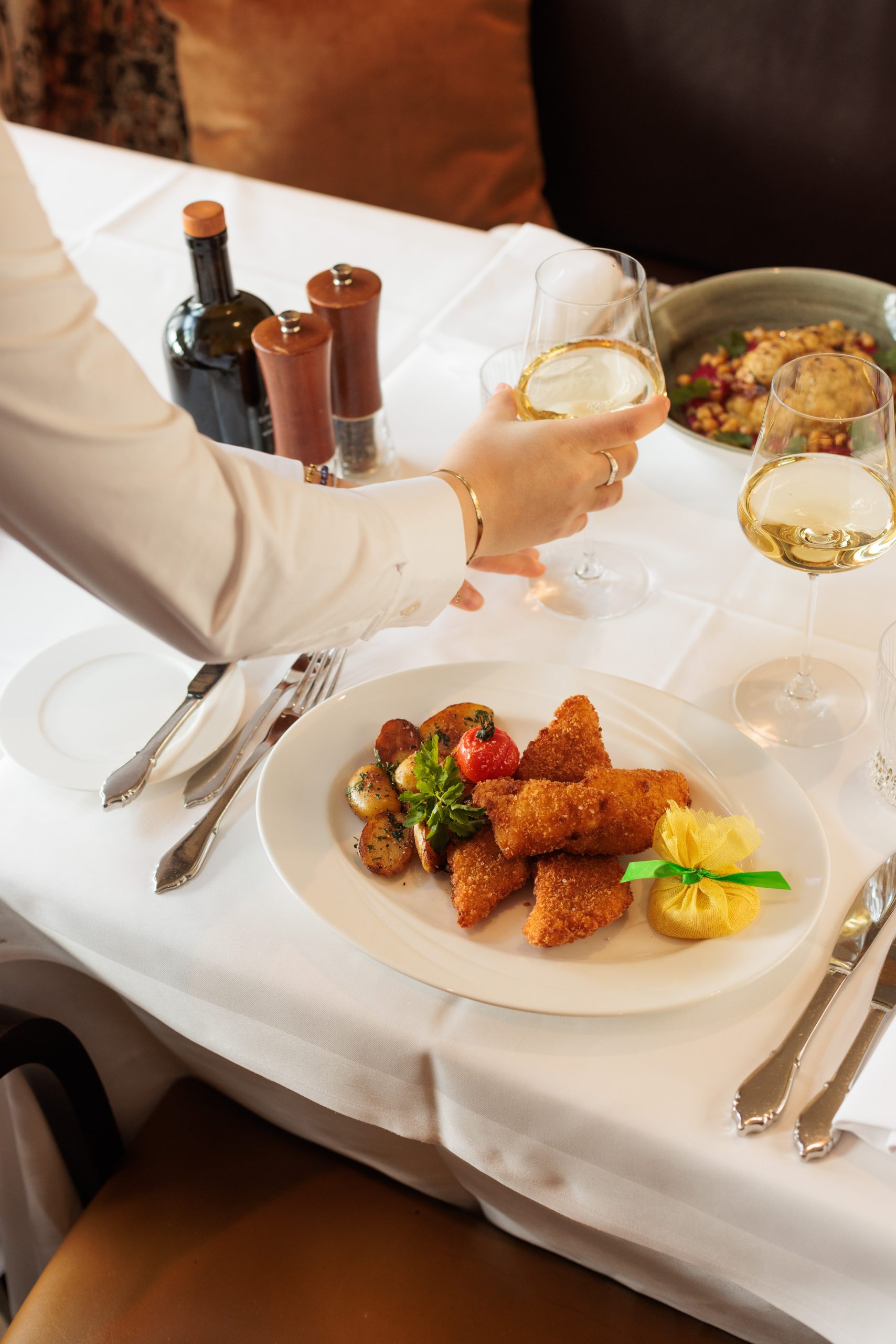 Eine Person mit einem Glas Weißwein sitzt an einem Tisch mit gebratenen Fischfilets, gebratenen Kartoffeln, Zitronenscheiben und Beilagen in der Sonnengallerie. Im Hintergrund sind ein zweites Weinglas, Gewürze und eine Schüssel mit Speisen zu sehen.