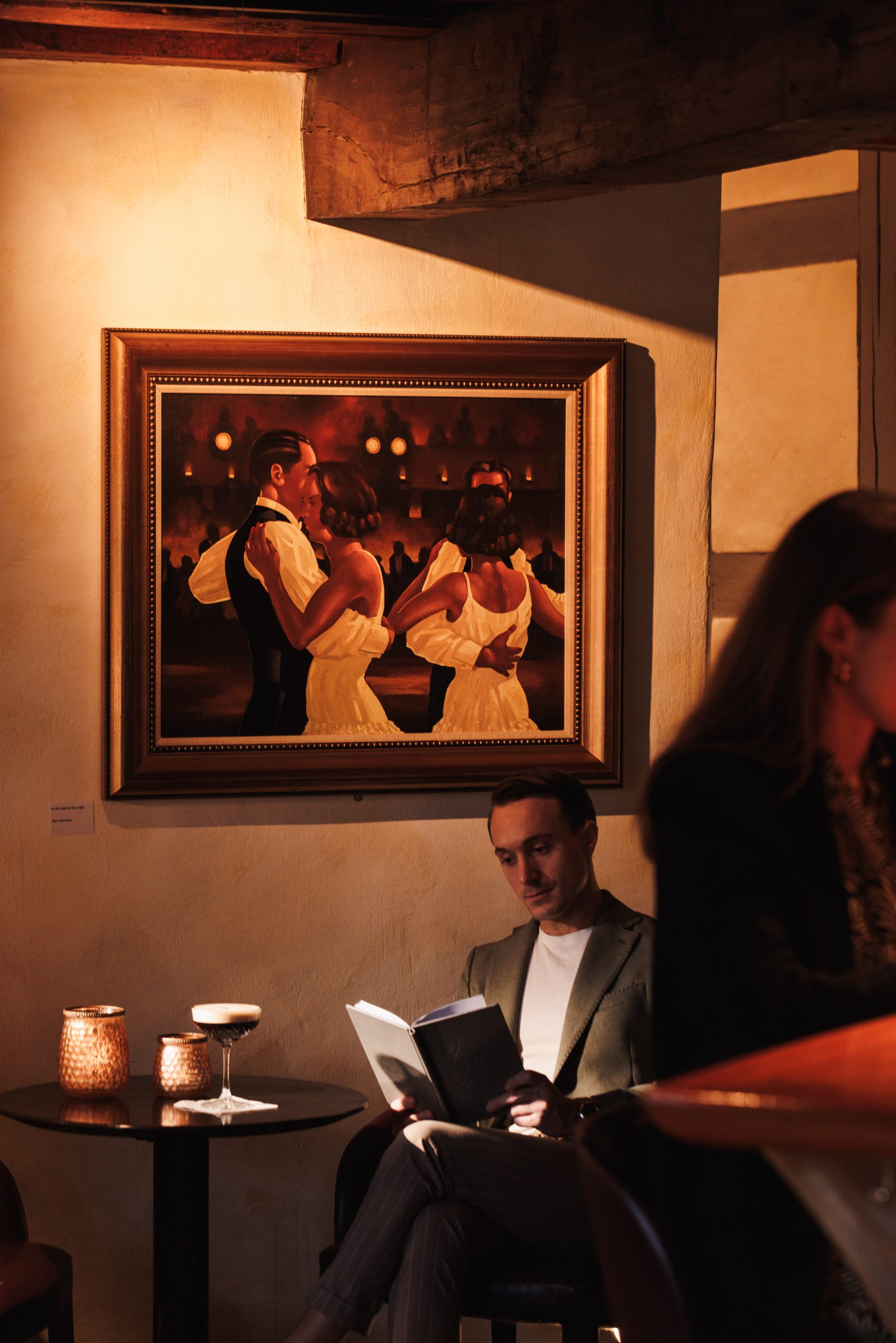 Ein Mann sitzt an einem schummrigen Tisch im Hotel Ambiente Küsnacht und liest neben zwei Cocktails und Kerzen ein Buch. Darüber hängt ein gerahmtes Gemälde mit tanzenden Paaren, während eine Frau in der warmen Atmosphäre neben der Menukarte an der Bar teilweise sichtbar ist.