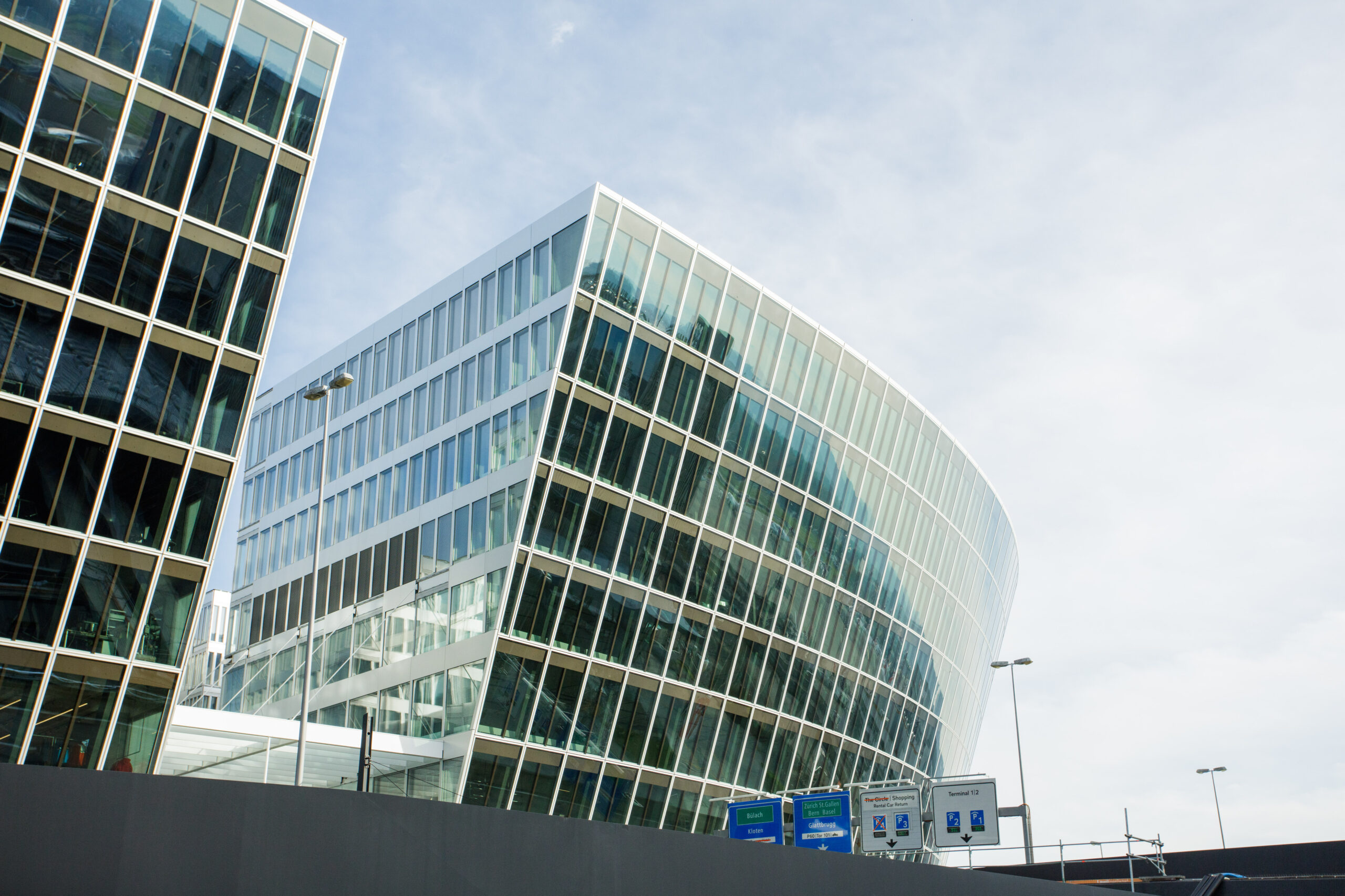 Ein modernes gläsernes Bürogebäude mit einer geschwungenen Fassade, in der sich der Himmel spiegelt, steht neben einem anderen kantigen Glasbau am Flughafen Zürich. Im Vordergrund sind bei teilweise bewölktem Himmel Straßenschilder und Laternenpfähle zu sehen.