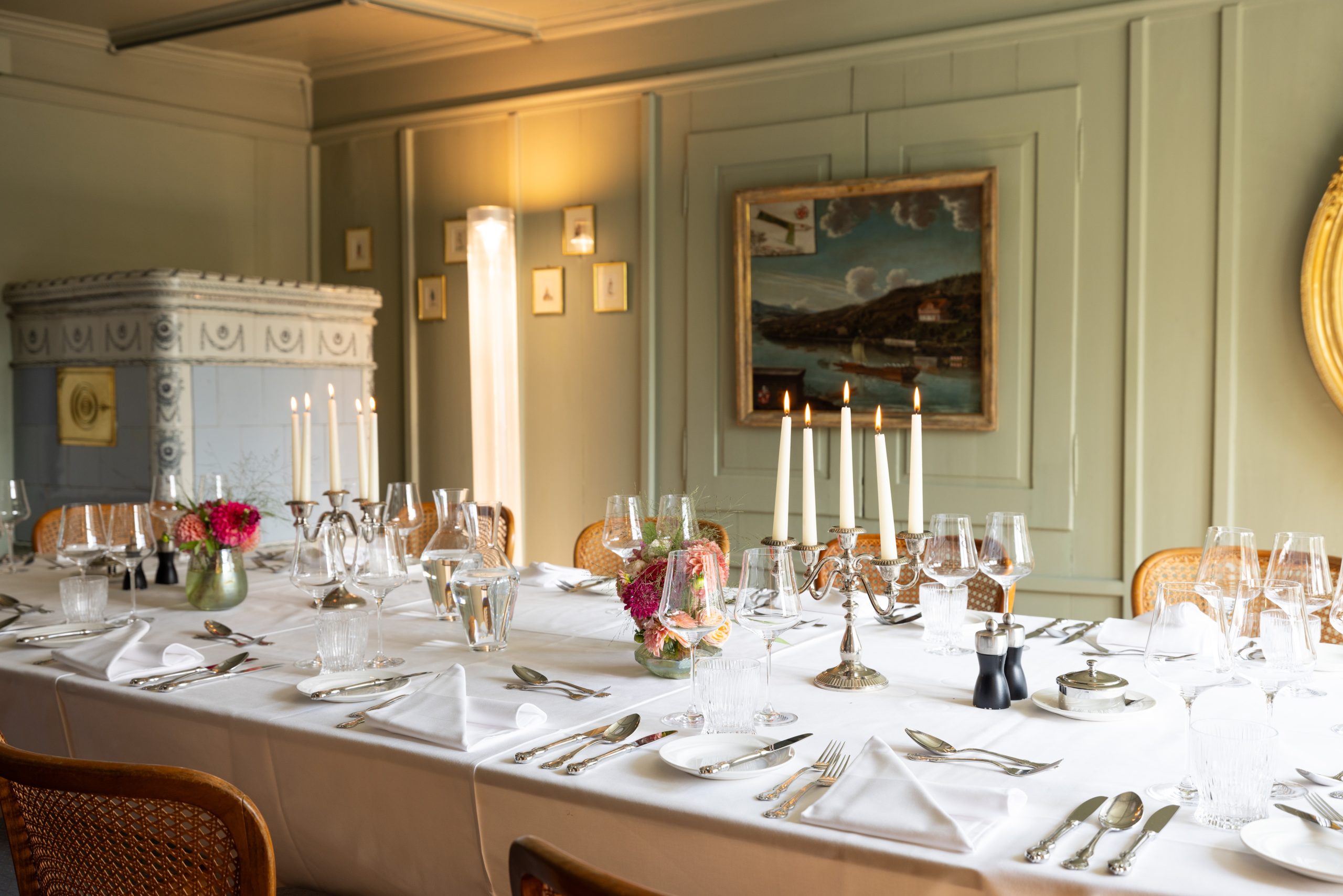 Ein formeller Esstisch mit weißer Bettwäsche, floralen Tafelaufsätzen, brennenden Kerzen, Gläsern und Silberbesteck schmückt die Prunkstube im Event Hotel Sonne mit hellgrünen Wänden, gerahmter Kunst und sanfter Beleuchtung.