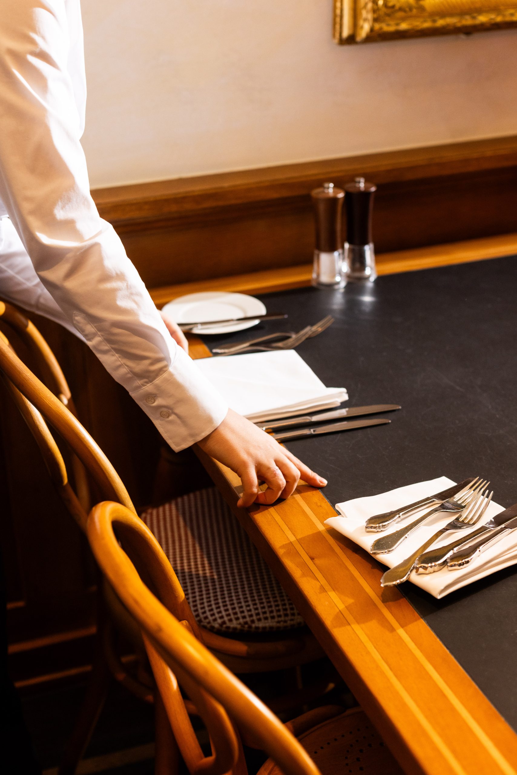 Eine Person in einem weißen Hemd deckt einen Holztisch mit Besteck, Servietten und Tellern im Hotel Sonne Gaststube, im Hintergrund sind Holzstühle und Salz- und Pfefferstreuer zu sehen.