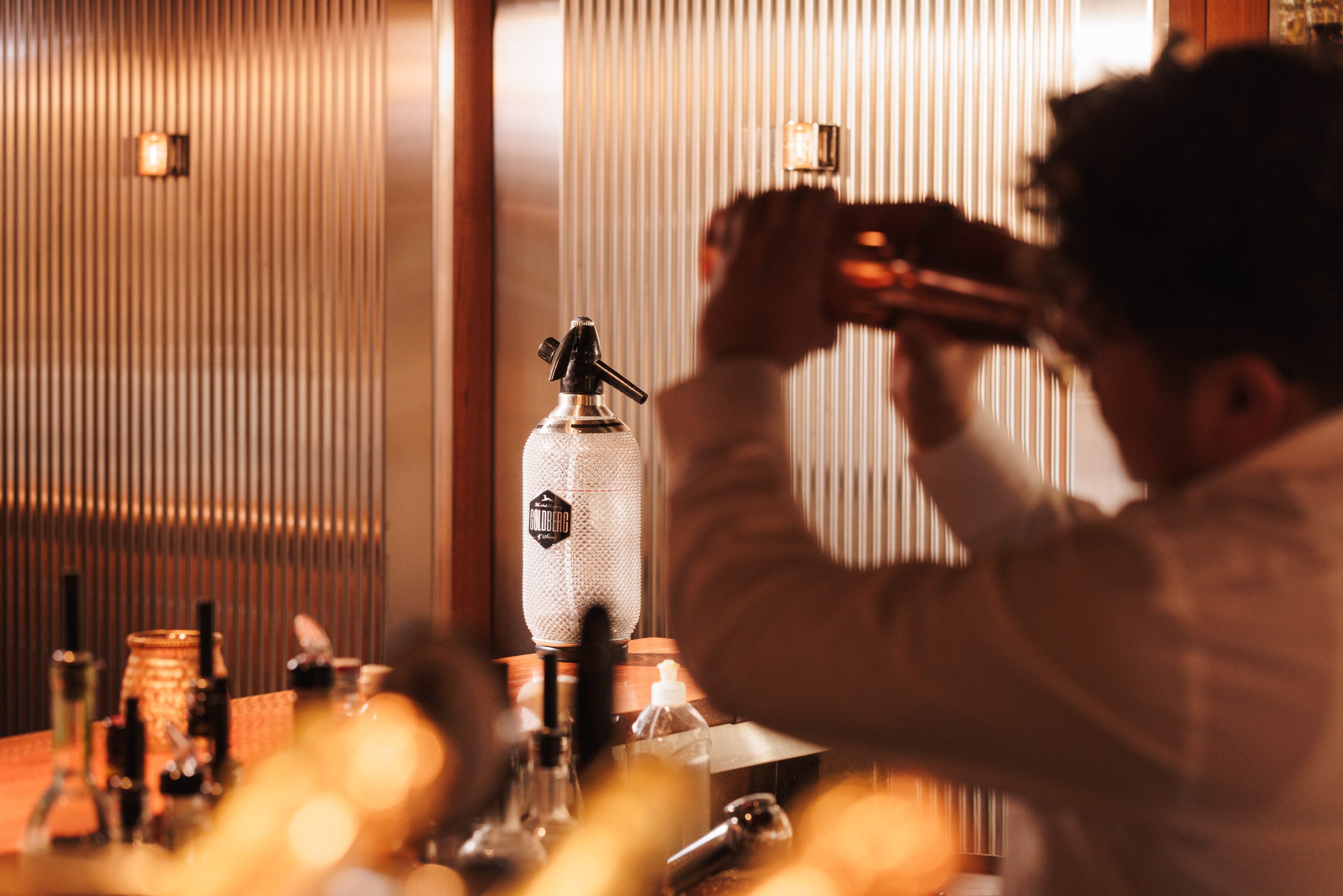 Ein Barkeeper im weißen Hemd schüttelt einen Cocktailshaker hinter der Turmbar im Hotel Sonne, wo Flaschen und ein Soda-Siphon auf der Arbeitsplatte in einem warm beleuchteten Ambiente stehen.