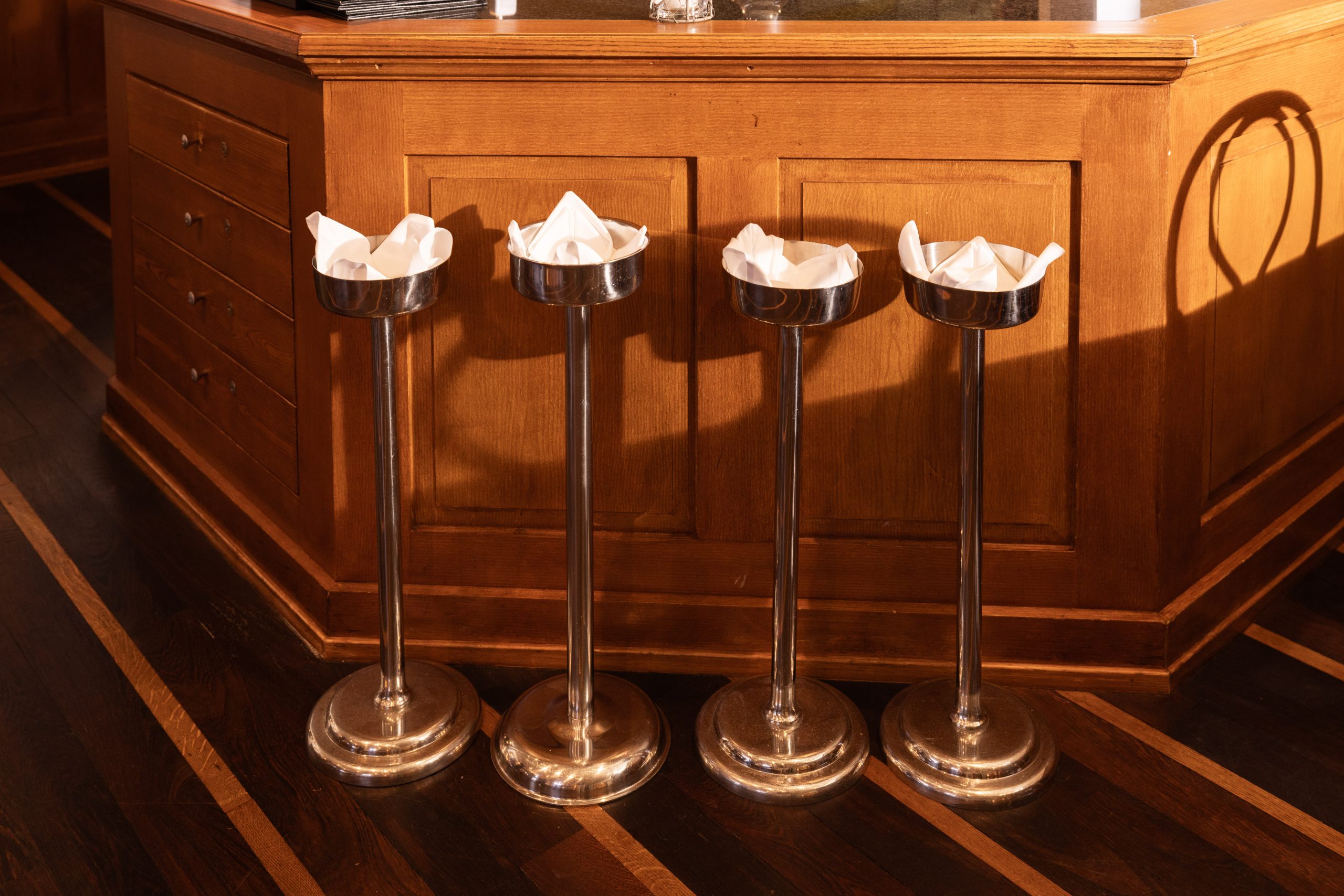 Vor dem hölzernen Tresen der Gaststube des Hotels Sonne stehen vier metallene Aschenbecher auf hohen Ständern. In jedem Aschenbecher liegen fein säuberlich gefaltete weiße Papierservietten, die vom warmen Sonnenlicht angestrahlt werden.