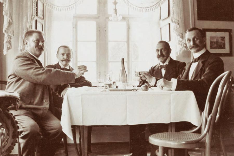 Vier Männer in Anzügen sitzen um einen kleinen Tisch mit Getränken in der hellen, im Vintage-Stil eingerichteten Lounge des Historie Hotel Sonne, stoßen an und unterhalten sich, während das Sonnenlicht durch die spitzenbesetzten Fenster hinter ihnen fällt.