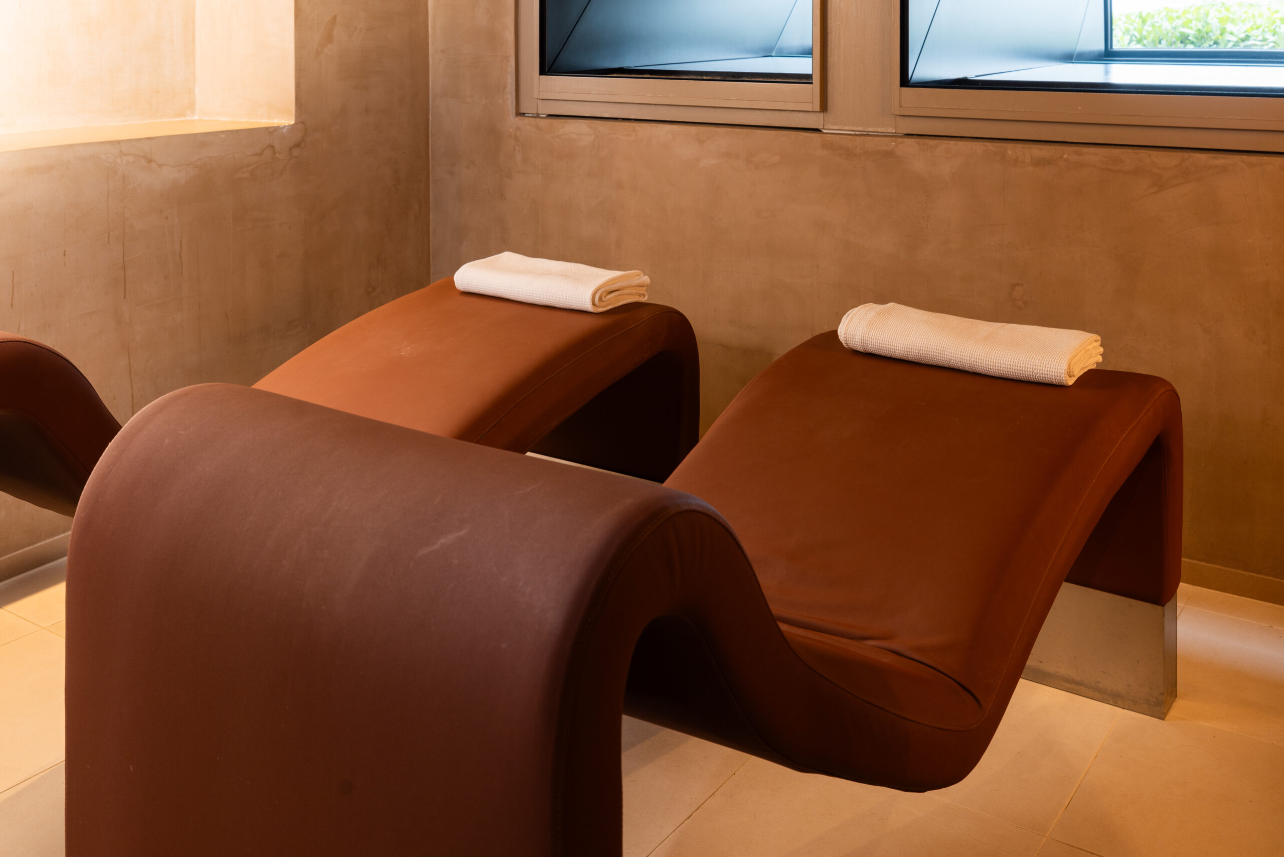 Zwei moderne, braune, wellenförmige Lounge-Sessel mit weißen, gerollten Handtüchern an den Kopfstützen stehen nebeneinander im sanft beleuchteten Wellness-Bereich des Hotel Sonne mit beigen Wänden und Fenstern.