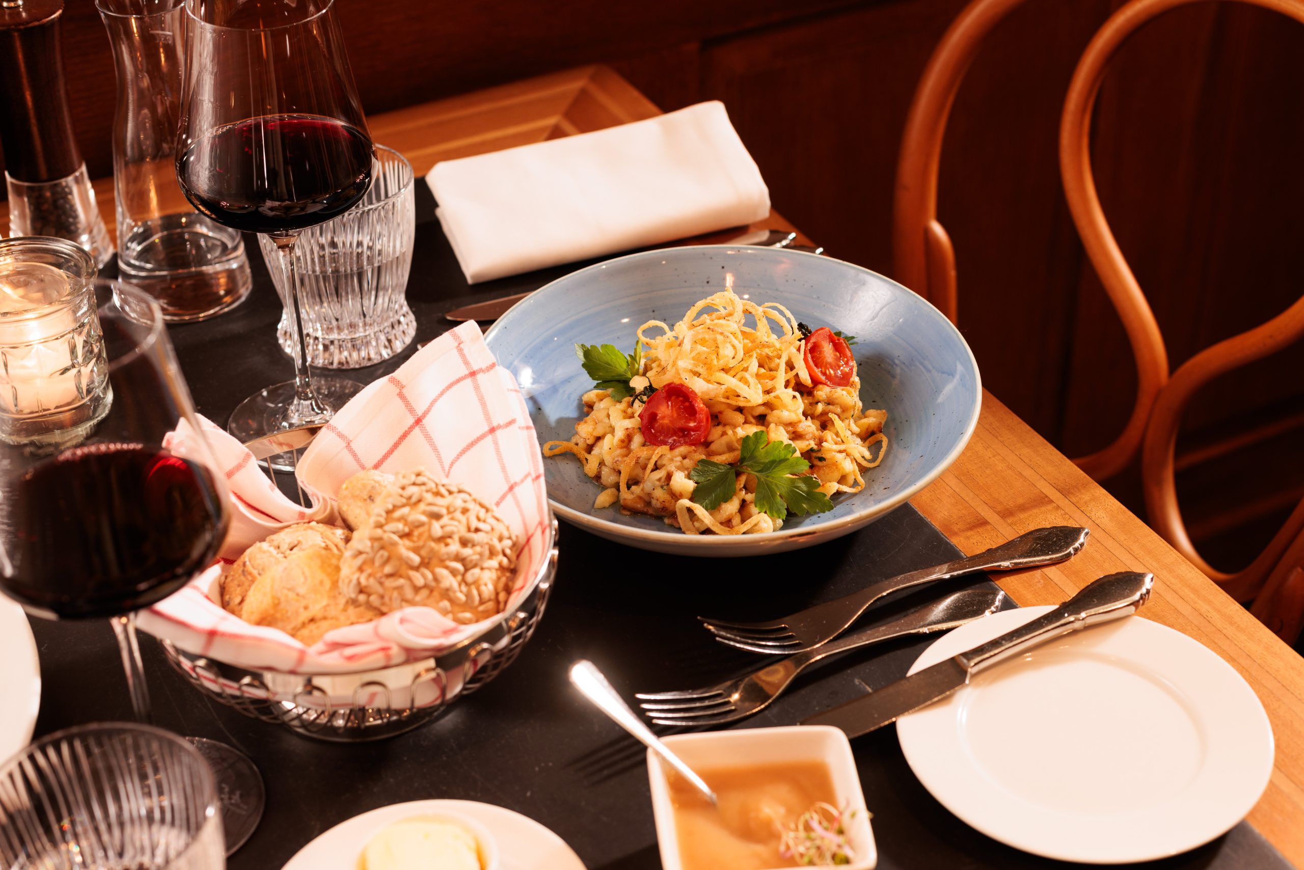 Ein gedeckter Tisch in der gemütlichen Gaststube des Hotels Sonne mit einer Schüssel Nudeln, garniert mit Kräutern und Kirschtomaten, einem Korb mit Brötchen, Gläsern mit Rotwein, Besteck, einer Serviette und einem Schälchen mit Butter.