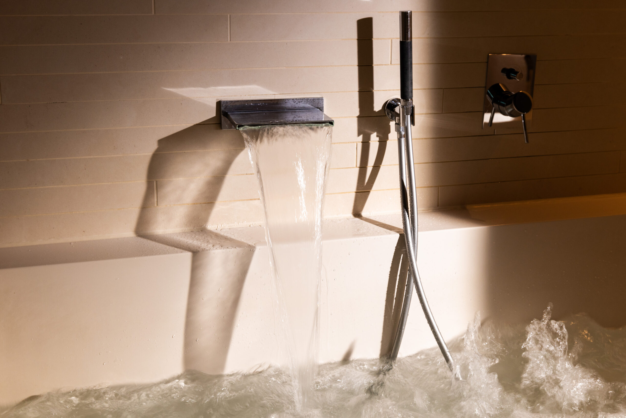 Eine moderne Badewanne im Hotel Sonne Küsnacht Spa füllt sich mit Wasser aus einem breiten, rechteckigen Auslauf an einer gefliesten Wand. In der Nähe befinden sich ein Handbrausekopf und Bedienknöpfe, während das Sonnenlicht weiche Schatten auf den Entspannungsraum wirft.