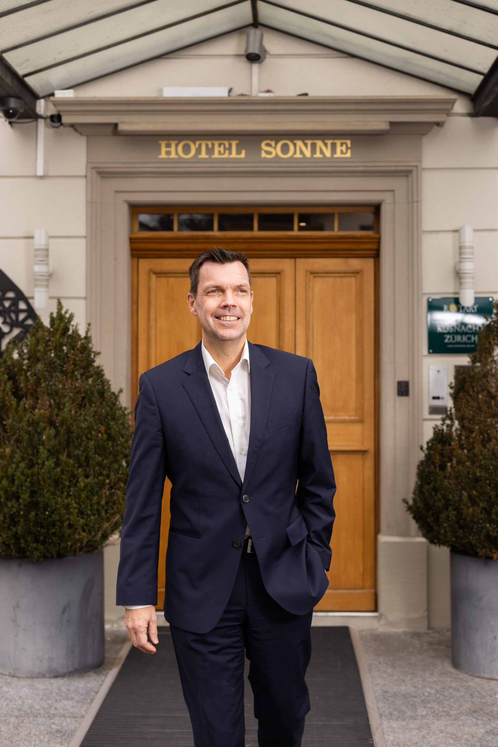 Harald Raab, im marineblauen Anzug, steht lächelnd vor dem Eingang des Hotels Sonne, umrahmt von großen Topfpflanzen auf beiden Seiten des Eingangs.
