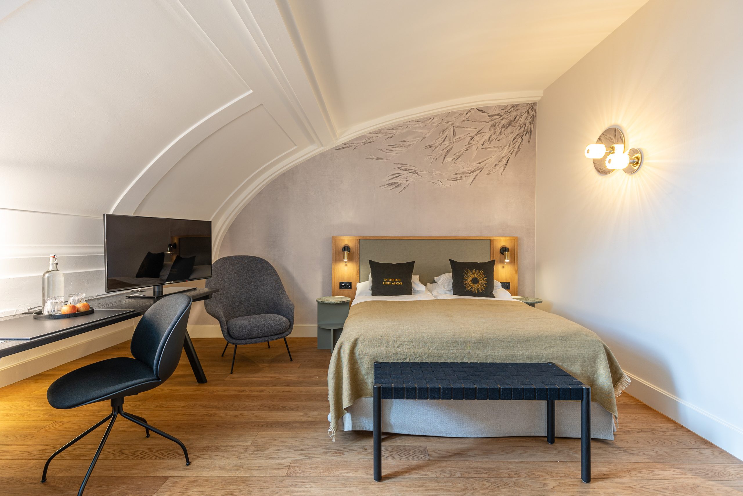 Ein modernes Hotelzimmer am See mit einem Doppelbett, hellem Bettzeug, zwei schwarzen Kissen, einem Wandbild, Schreibtisch und Monitor, Stuhl, Sessel, Holzboden und Wandleuchten. Das Zimmer verfügt über eine gebogene Decke für zusätzlichen Stil.