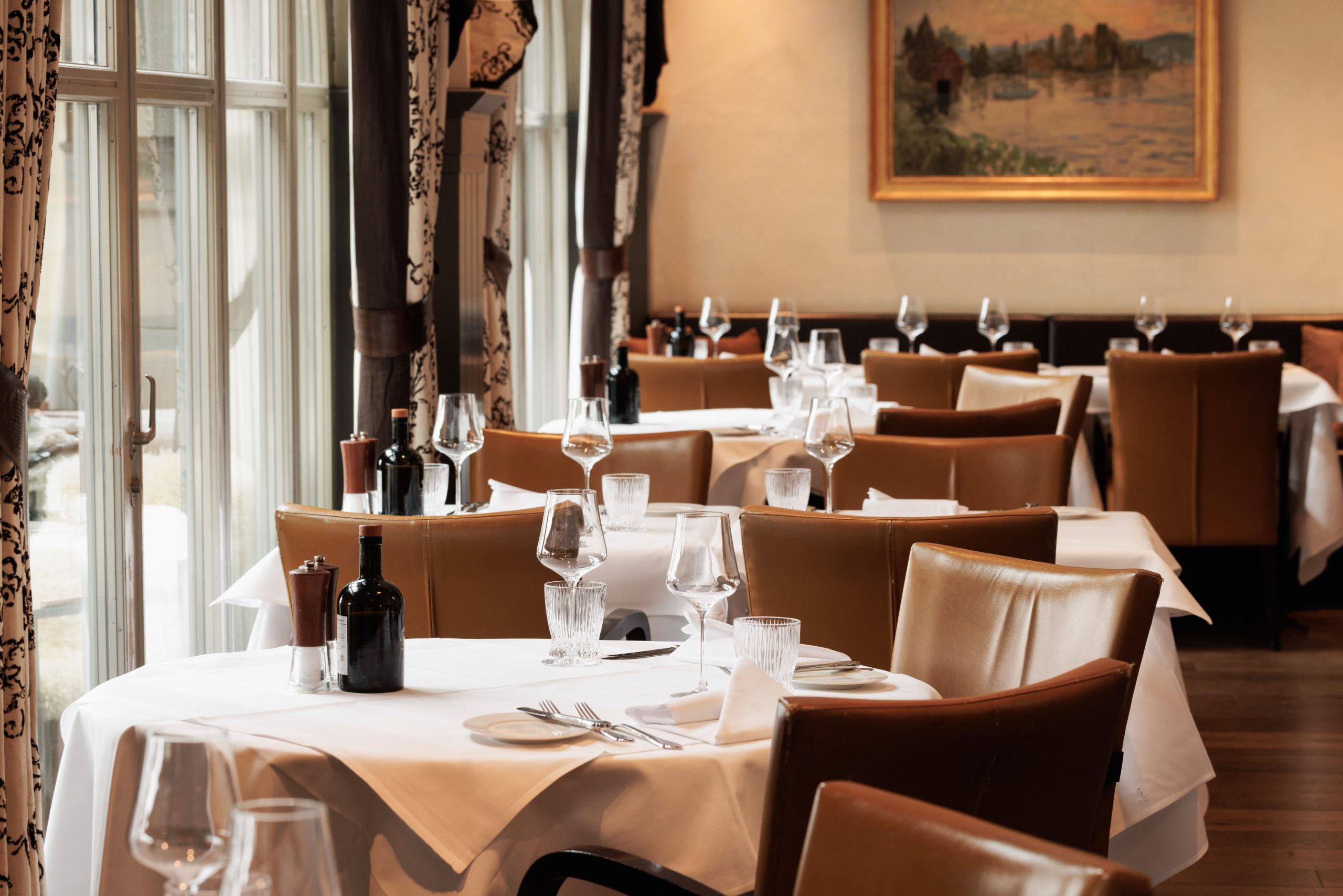 Elegantes Restaurant Sonne mit ordentlich gedeckten Tischen mit weißen Tüchern, braunen Lederstühlen und funkelnden Weingläsern. Große Fenster und Wandkunst schaffen eine warme Atmosphäre. Es sind keine Menschen anwesend.