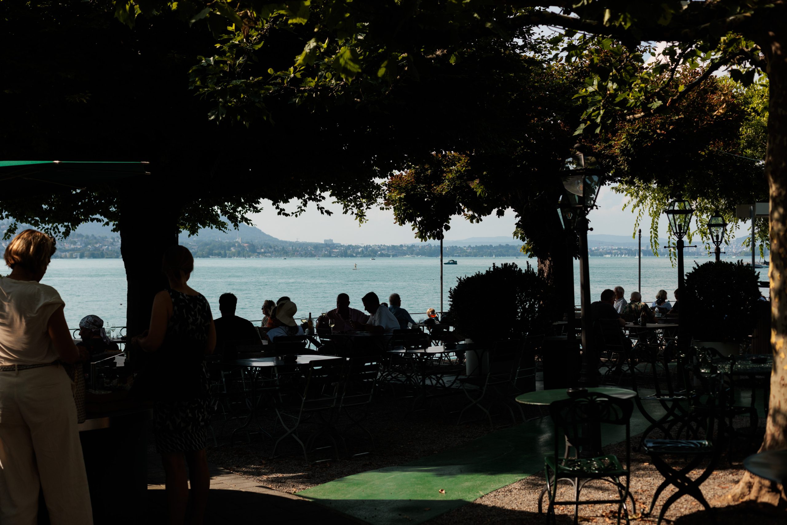 Die Menschen genießen das Frühstück an schattigen Cafétischen im Freien am See, umgeben von grünen Bäumen. Die warme Sonne glitzert auf dem Wasser, und die fernen Berge unter dem hellen Himmel schaffen eine entspannte und gesellige Atmosphäre.