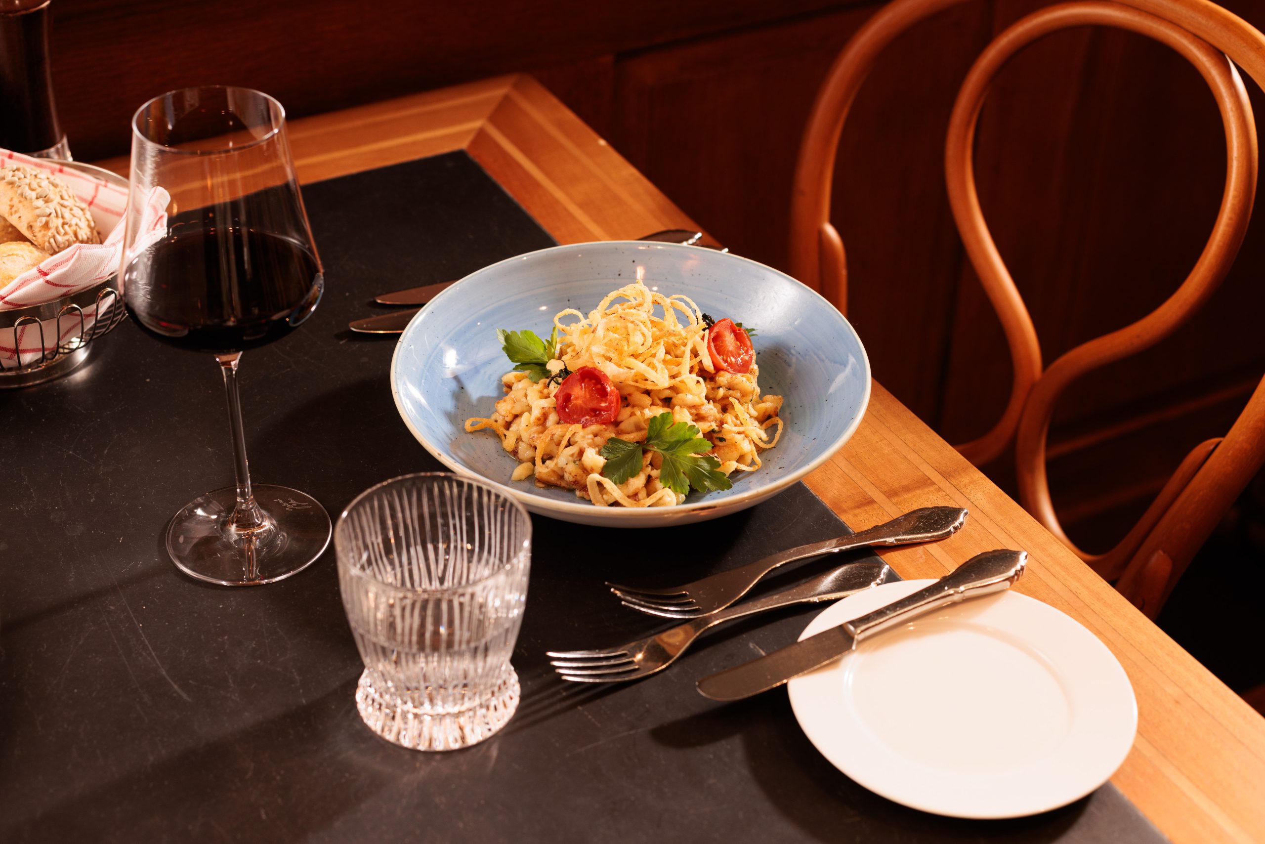 In der gemütlichen Gaststube des Hotels Sonne steht eine blaue Schüssel mit Nudeln, garniert mit Tomaten, Petersilie und knusprigen Zwiebeln, auf einem dunklen Tisch neben Rotwein, Wasser, Besteck, einem leeren Teller und einem Korb mit Brot.