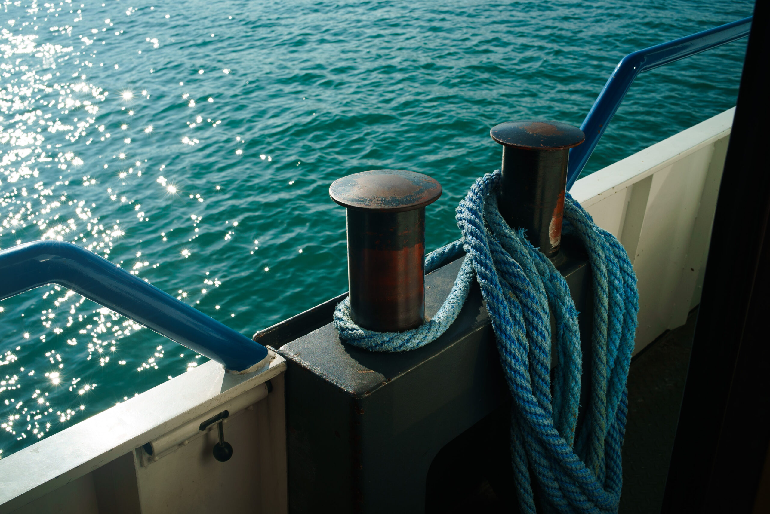 Nahaufnahme eines blauen Seils, das um einen Metallpoller auf einem Boot gewickelt ist. Im Hintergrund spiegelt sich das Sonnenlicht auf dem ruhigen, türkisfarbenen Wasser des Küsnachter Schiffes Anlege.
