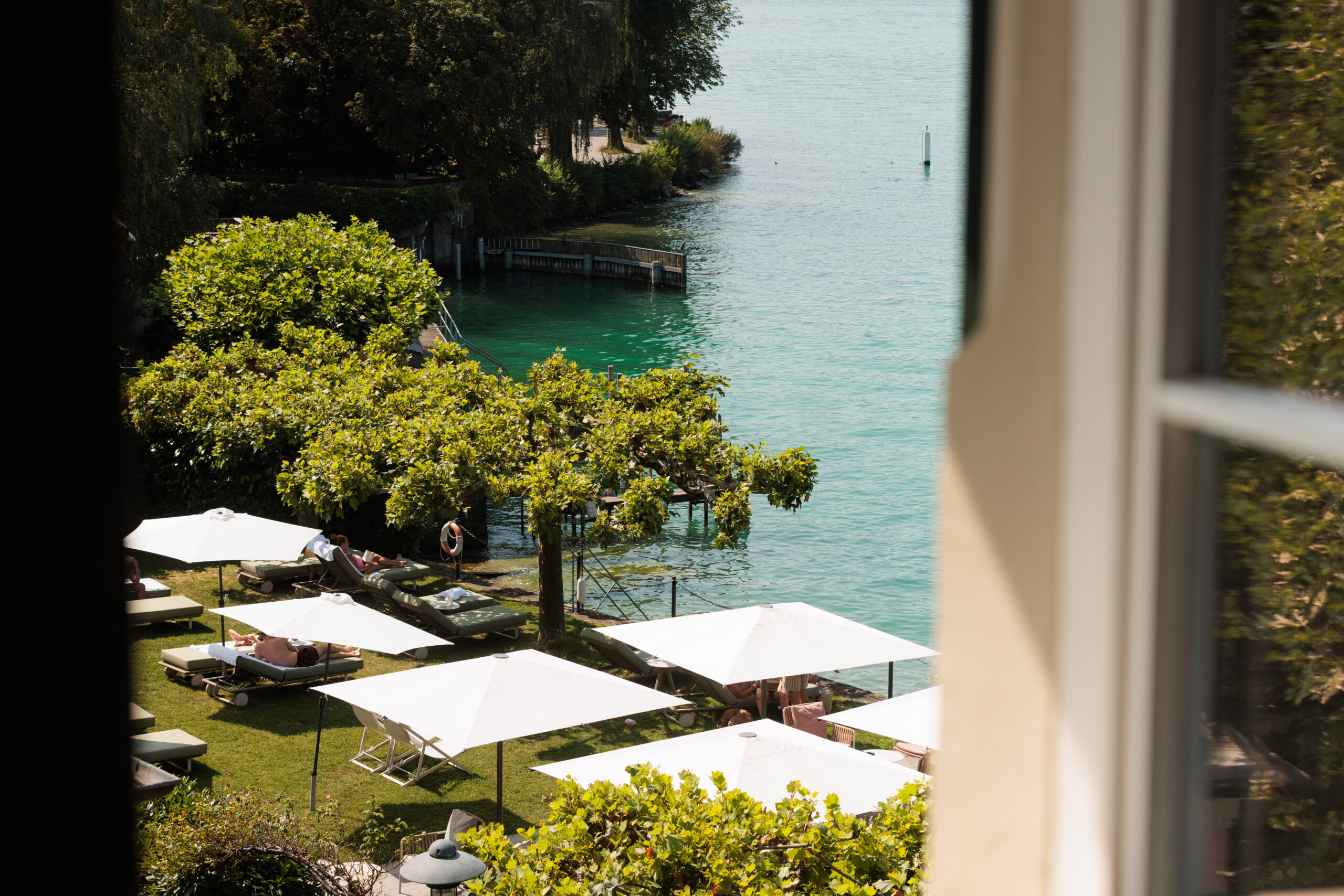 Geniessen Sie die Aussicht vom Hotel Sonne, mit Blick auf einen ruhigen Seegarten am Zürichsee, mit weissen Sonnenschirmen, Liegestühlen und Menschen, die sich an einem sonnigen Tag unter grünen Bäumen entspannen.