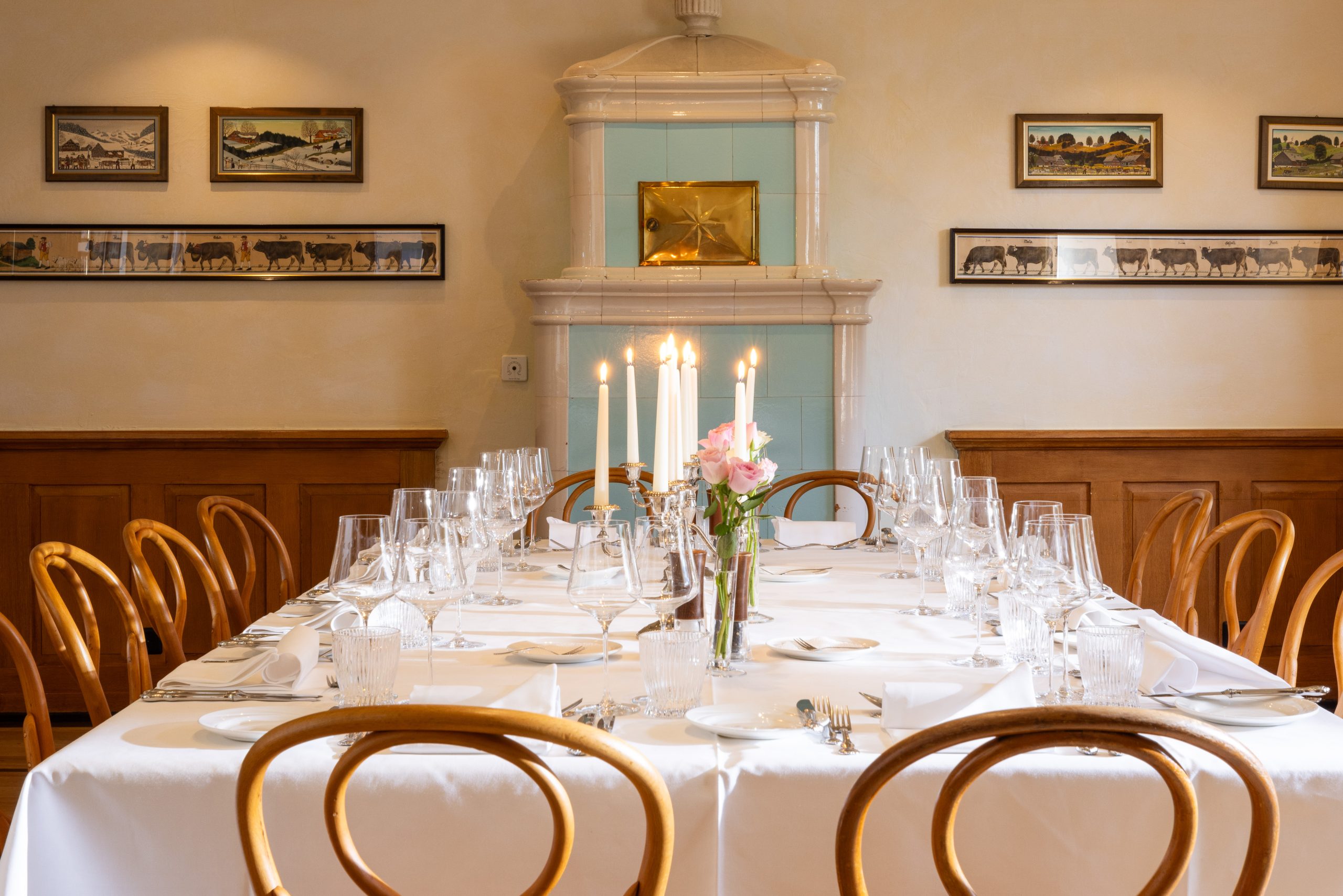 Elegantes Esszimmer Eventraum mit einem quadratischen Tisch, der für eine Mahlzeit gedeckt ist, mit weißen Tischtüchern, Holzstühlen, Kerzen und rosa Blumen; klassisches Dekor mit gerahmter Kunst und einem Kachelofen - perfekt für diejenigen, die eine Location mieten möchten.