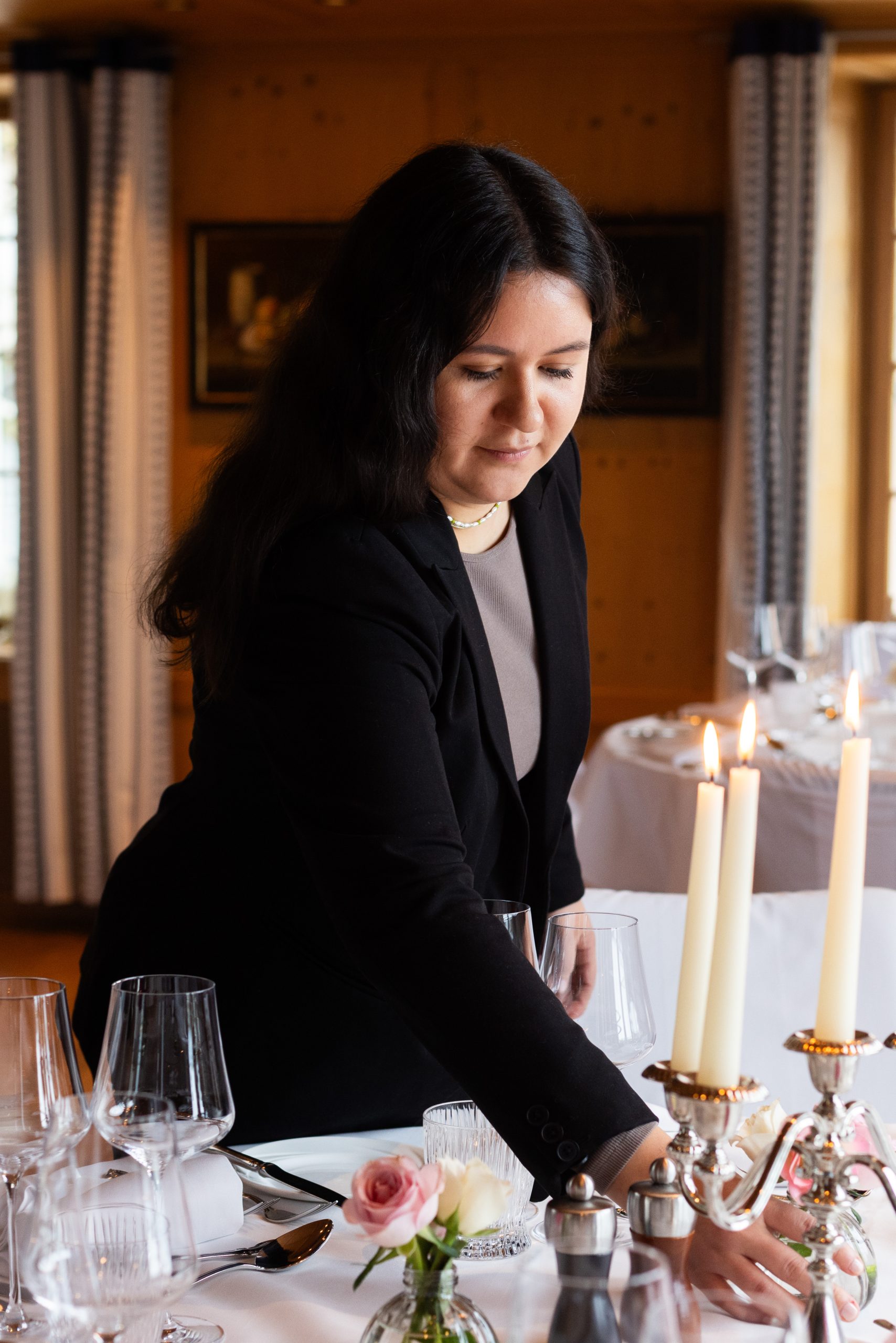 Eine Frau mit langen dunklen Haaren und einem Blazer deckt einen elegant gedeckten Tisch mit Weingläsern, Kerzen und einer rosafarbenen Rose in einem gedämpft beleuchteten Esszimmer.