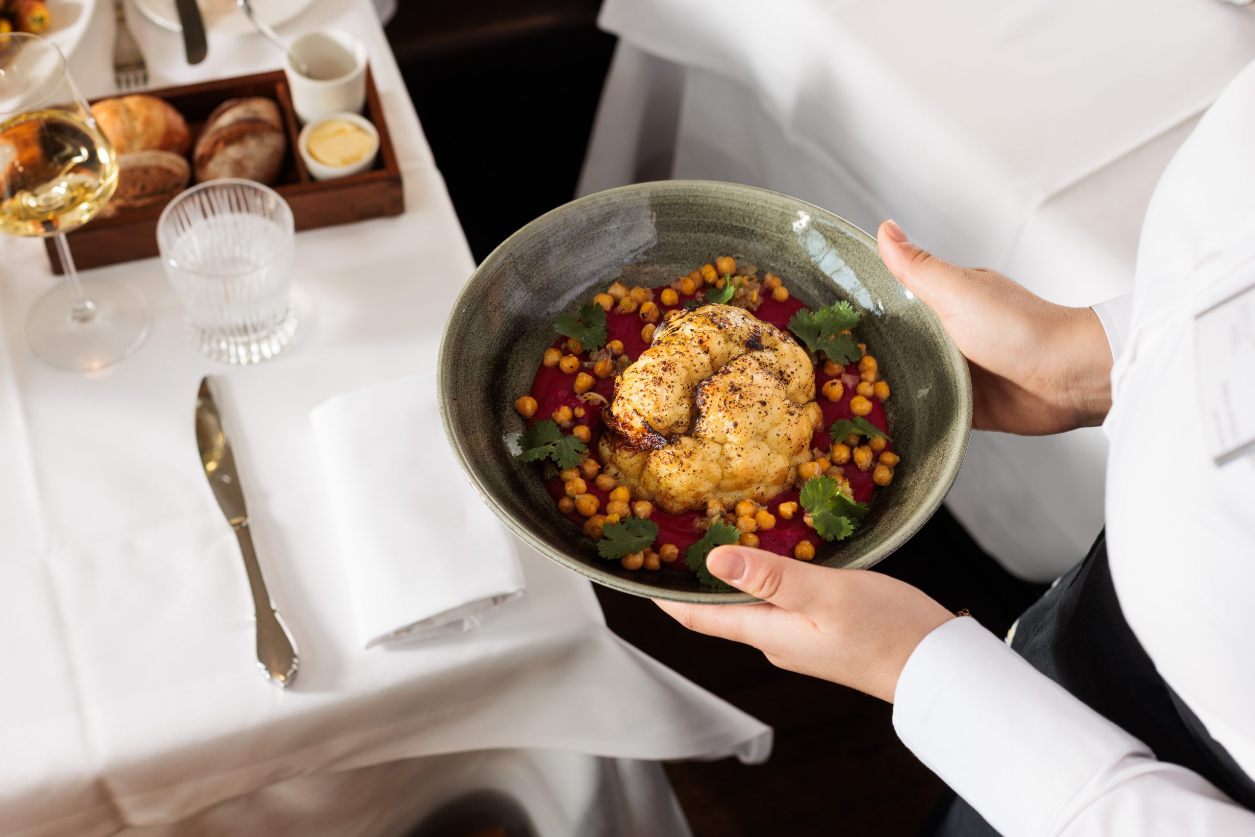 Eine Person in einem weißen Hemd hält eine grüne Schale mit gebratenem Blumenkohl, Kichererbsen, Tomatensauce und Kräutern - ein farbenfrohes Sonnengalerie Gericht. Der Tisch ist mit Brot, Wein, einem Glas und Besteck auf weißen Tischtüchern gedeckt.