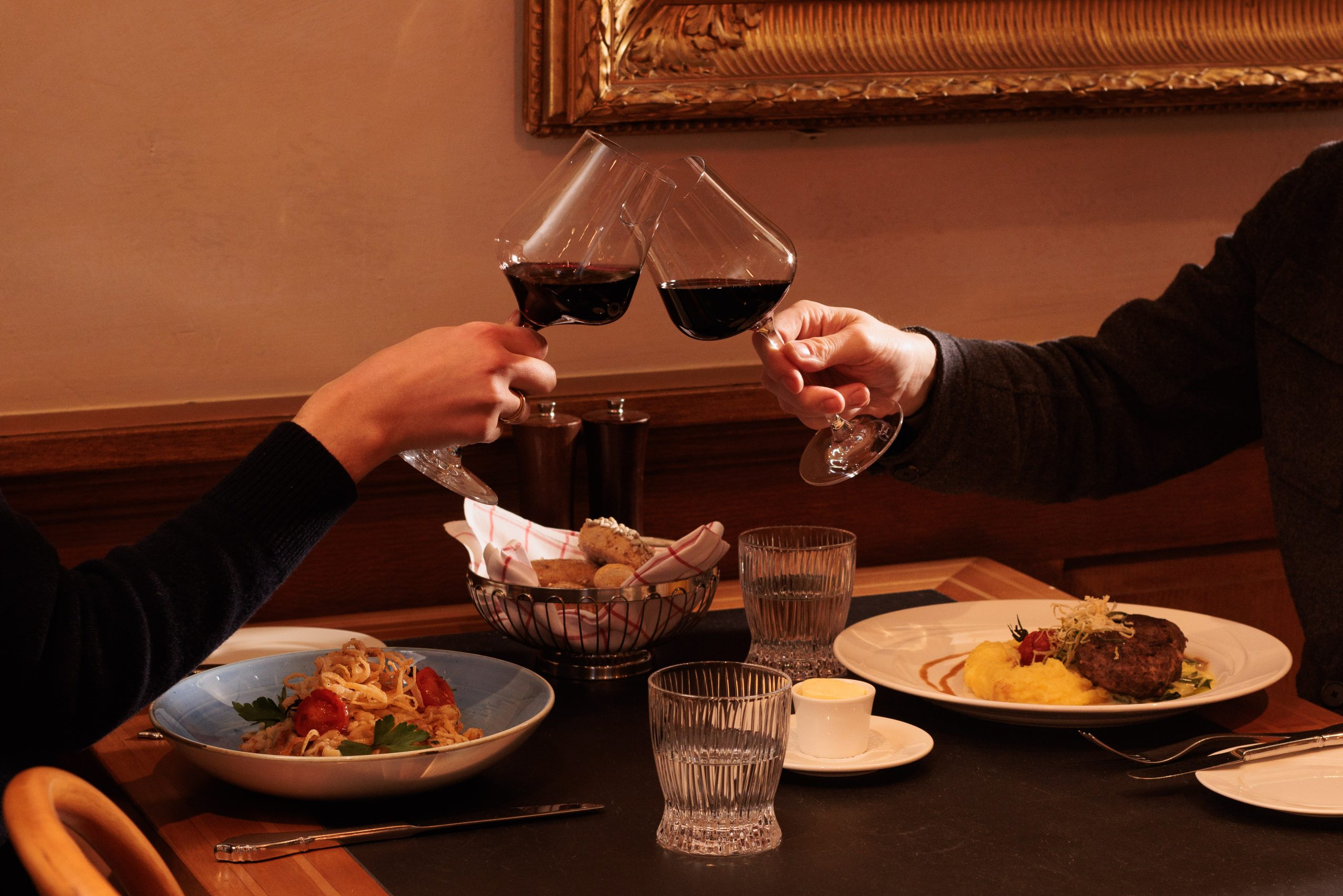 Zwei Personen stoßen mit Rotwein an einem Tisch in der Gaststube des Hotels Sonne an, der mit Tellern mit Nudeln, Steak, Kartoffelpüree, Brot und Wassergläsern gedeckt ist, und schaffen so eine warme und elegante Essatmosphäre.