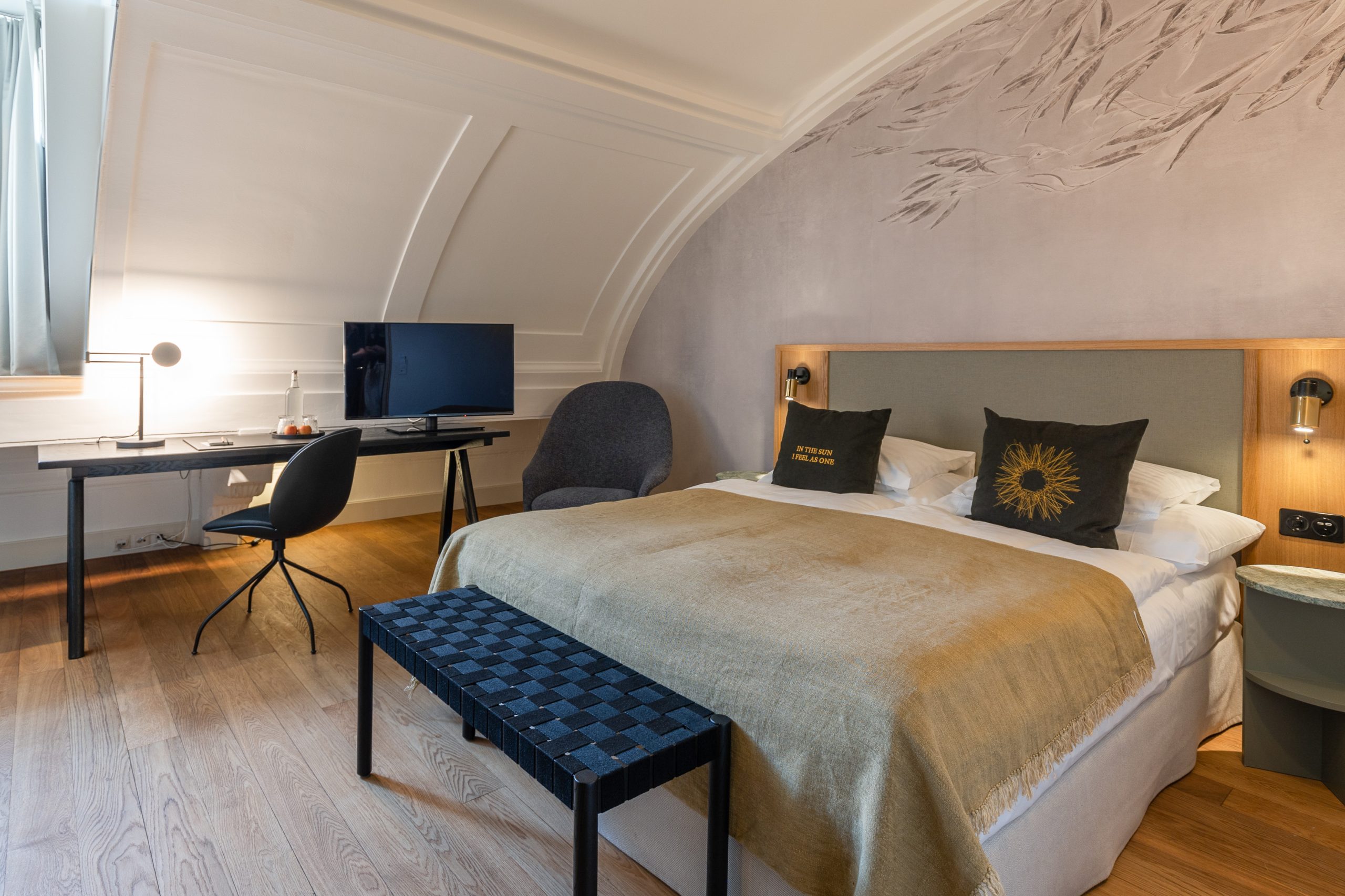 Ein modernes Zimmer im Boutiquehotel Sonne mit einem Doppelbett, zwei Kissen, einer gemusterten Bank am Fußende des Bettes, einem Schreibtisch mit Stuhl, einem Fernseher, einer Lampe und einem grauen Sessel. Der Raum verfügt über Holzböden und eine dekorative Akzentwand.