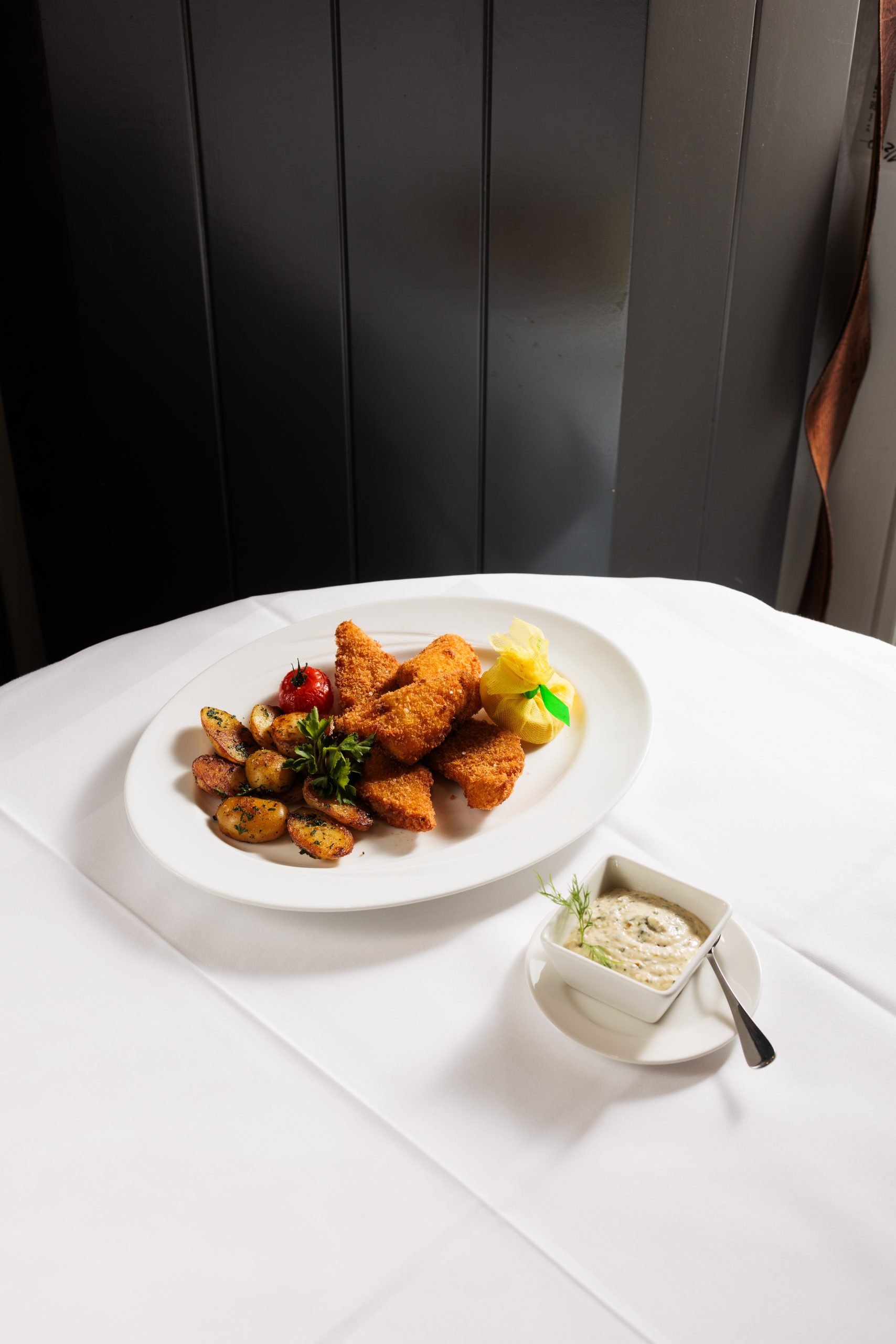 In der Sonnengalerie steht auf einem weißen Tischtuch ein weißer Teller mit paniertem Bratfisch, Röstkartoffeln, einer Zitronenspalte und Garnitur, daneben ein Schälchen mit Sahnesoße mit einem Löffel und einem Dillzweig.