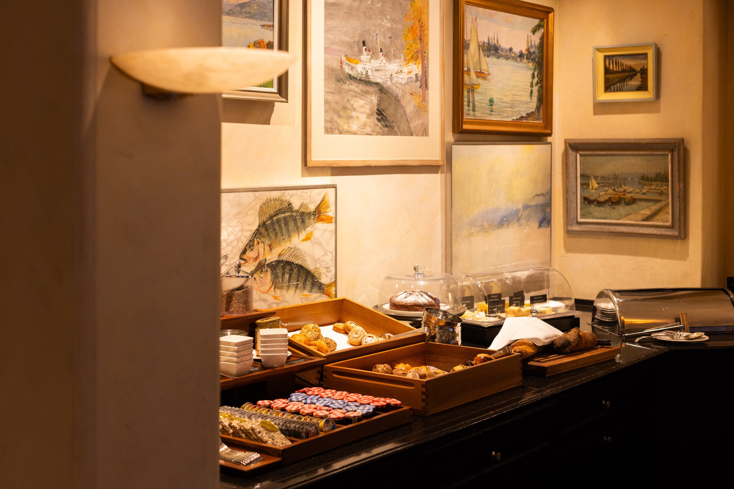 A buffet setup with pastries, bread, and spreads in wooden trays on a counter, framed by paintings of boats and fish on a warmly-lit wall in the background.
