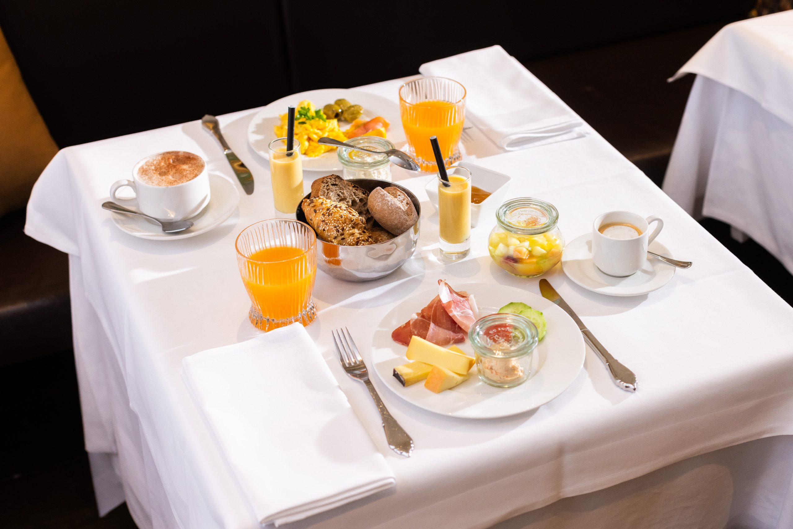 A neatly set breakfast table for two with juice, coffee, assorted breads, sliced meats, cheese, fruit cups, and smoothies on white tablecloths.