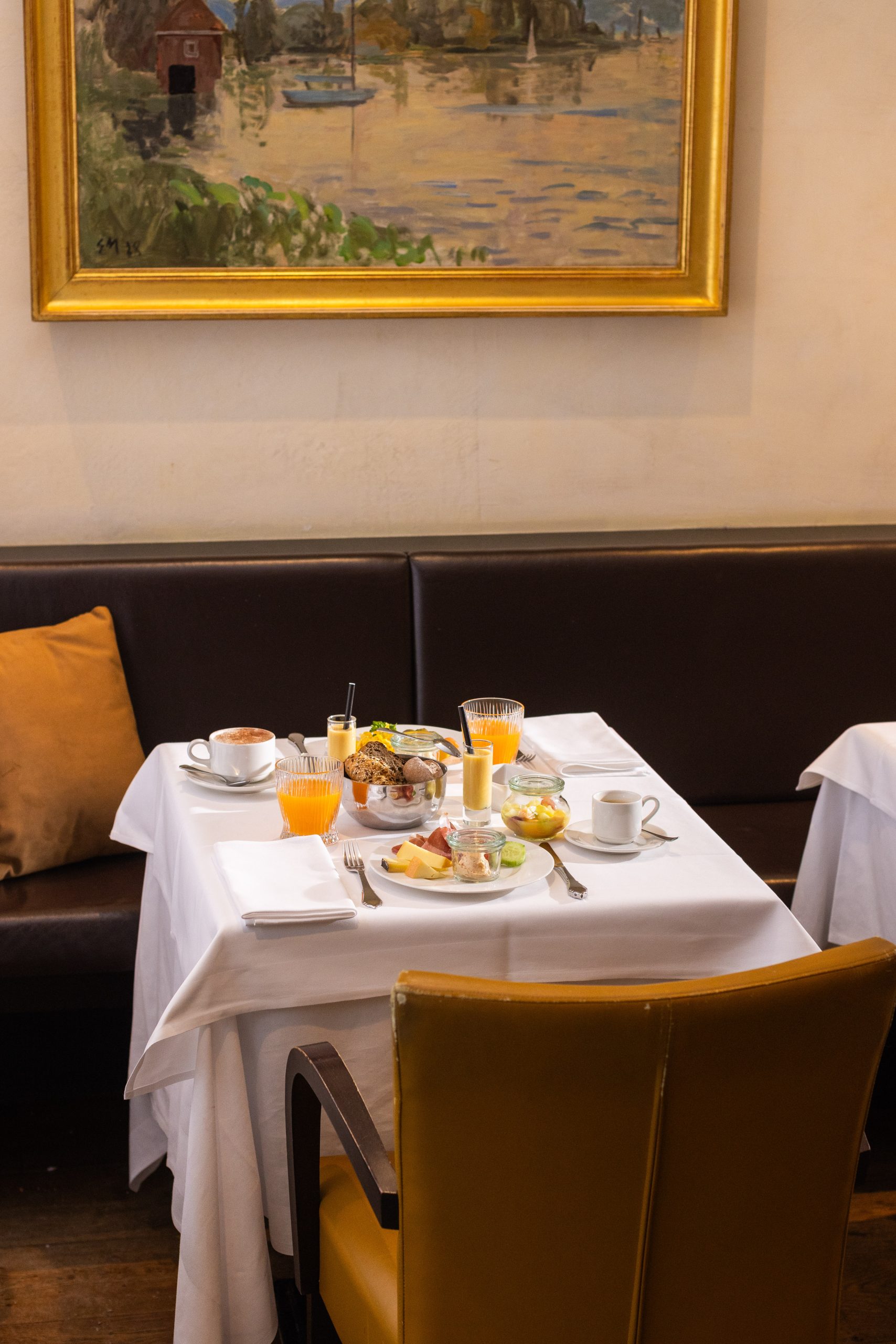 A small dining table set for two with breakfast foods, juice, coffee, and pastries. The table is covered with a white cloth and surrounded by brown and yellow chairs. A large framed painting hangs on the wall behind.