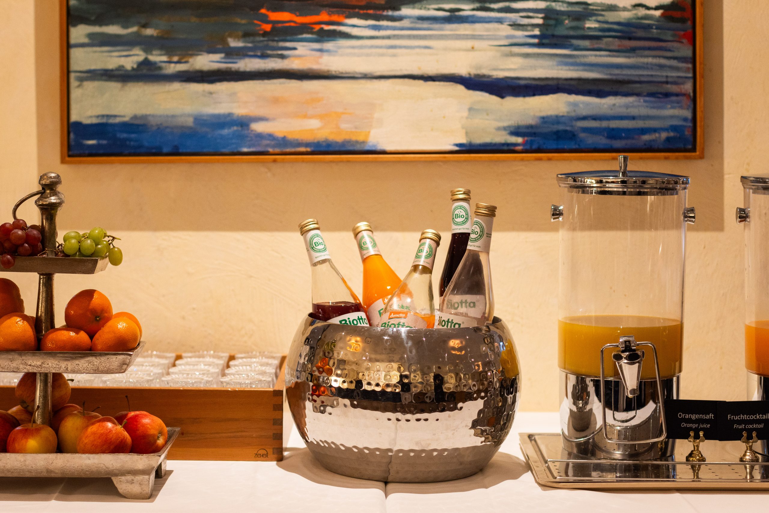 A breakfast buffet setup with a tiered tray of apples and grapes, bottles of drinks in a silver ice bucket, a wooden box of glasses, and a dispenser filled with orange juice. A colorful painting hangs on the wall behind.