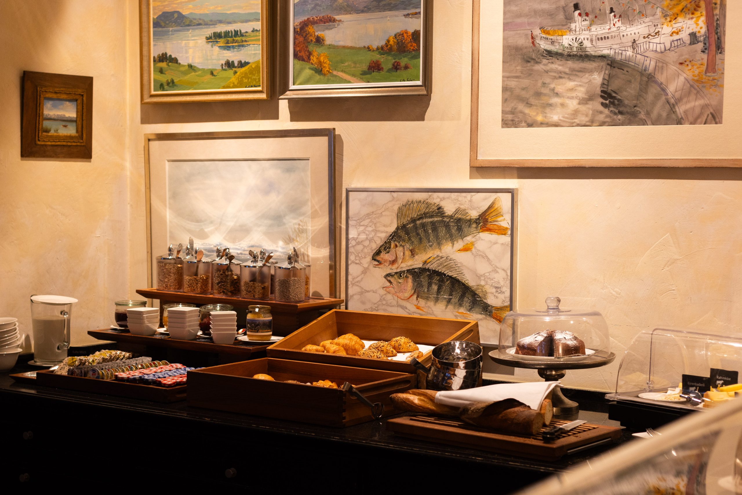 A breakfast buffet with pastries, bread, cereals, and cakes is set on a counter under framed paintings, including landscapes and fish. Plates, cups, and utensils are neatly arranged to the side.