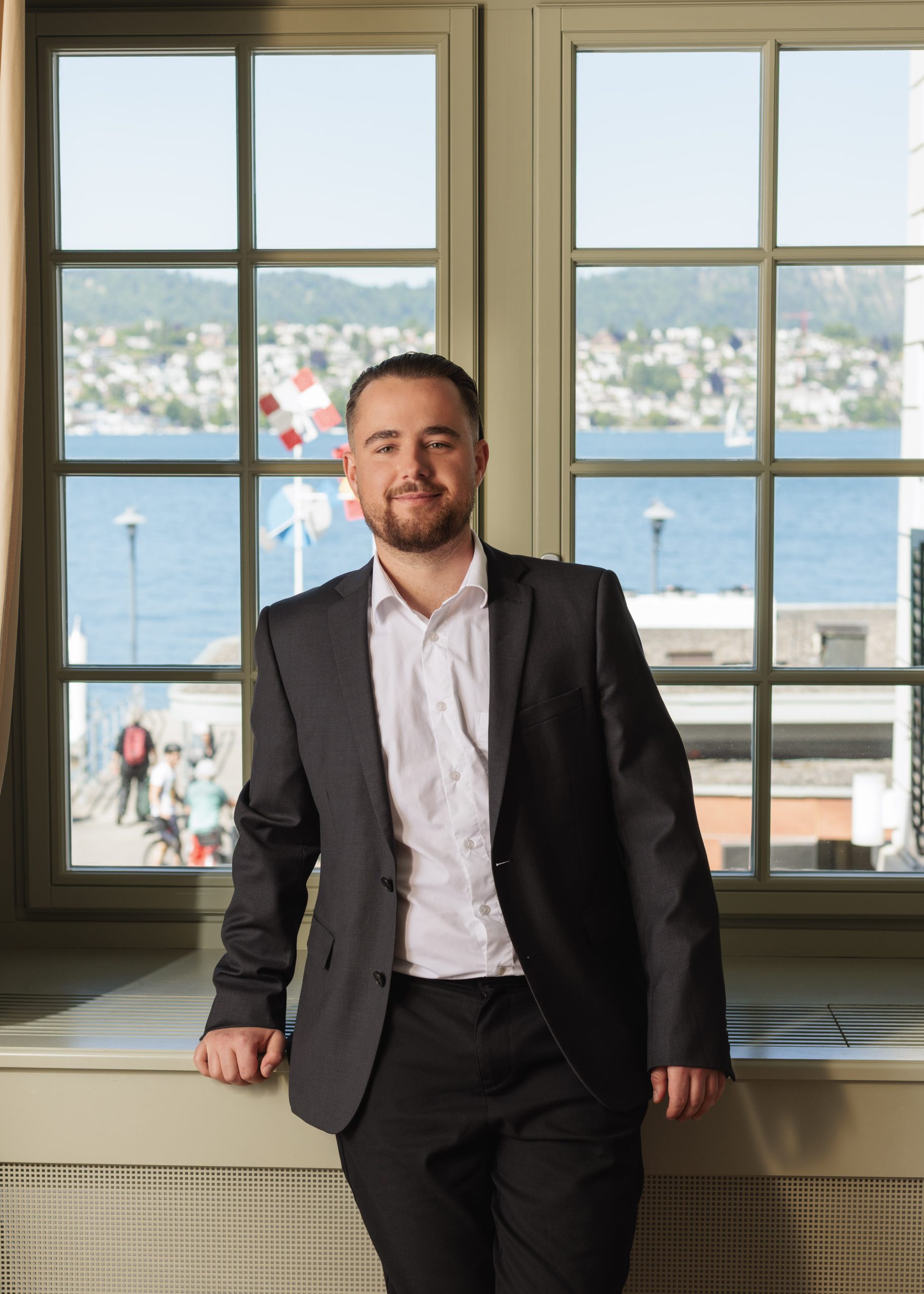 Ein Mann im Anzug steht drinnen an einem großen Fenster mit Blick auf eine Stadt am See und Schweizer Fahnen draußen. Er lächelt und schaut in die Kamera.
