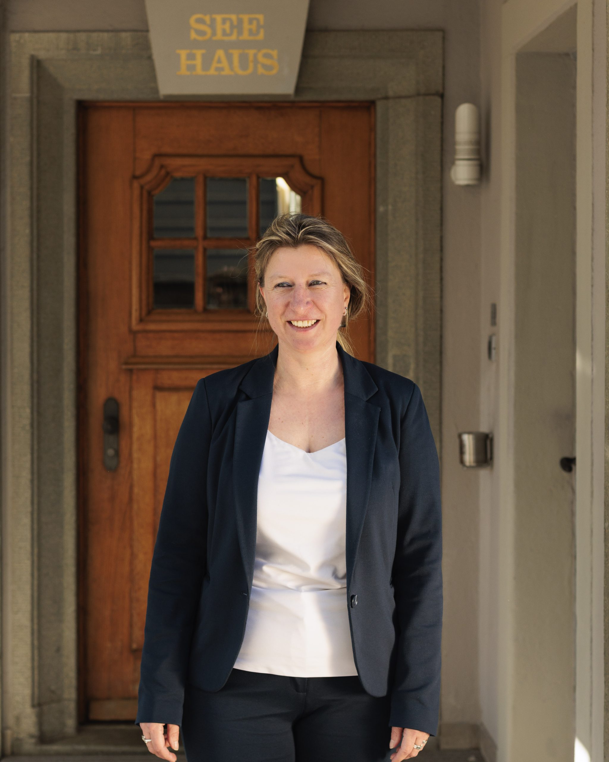 Eine Frau mit blondem Haar, die einen marineblauen Blazer und ein weißes Oberteil trägt, steht lächelnd vor einer Holztür. Über der Tür steht ein Schild mit der Aufschrift SEE HAUS. Der Schauplatz scheint ein Eingang zu einem Gebäude zu sein.