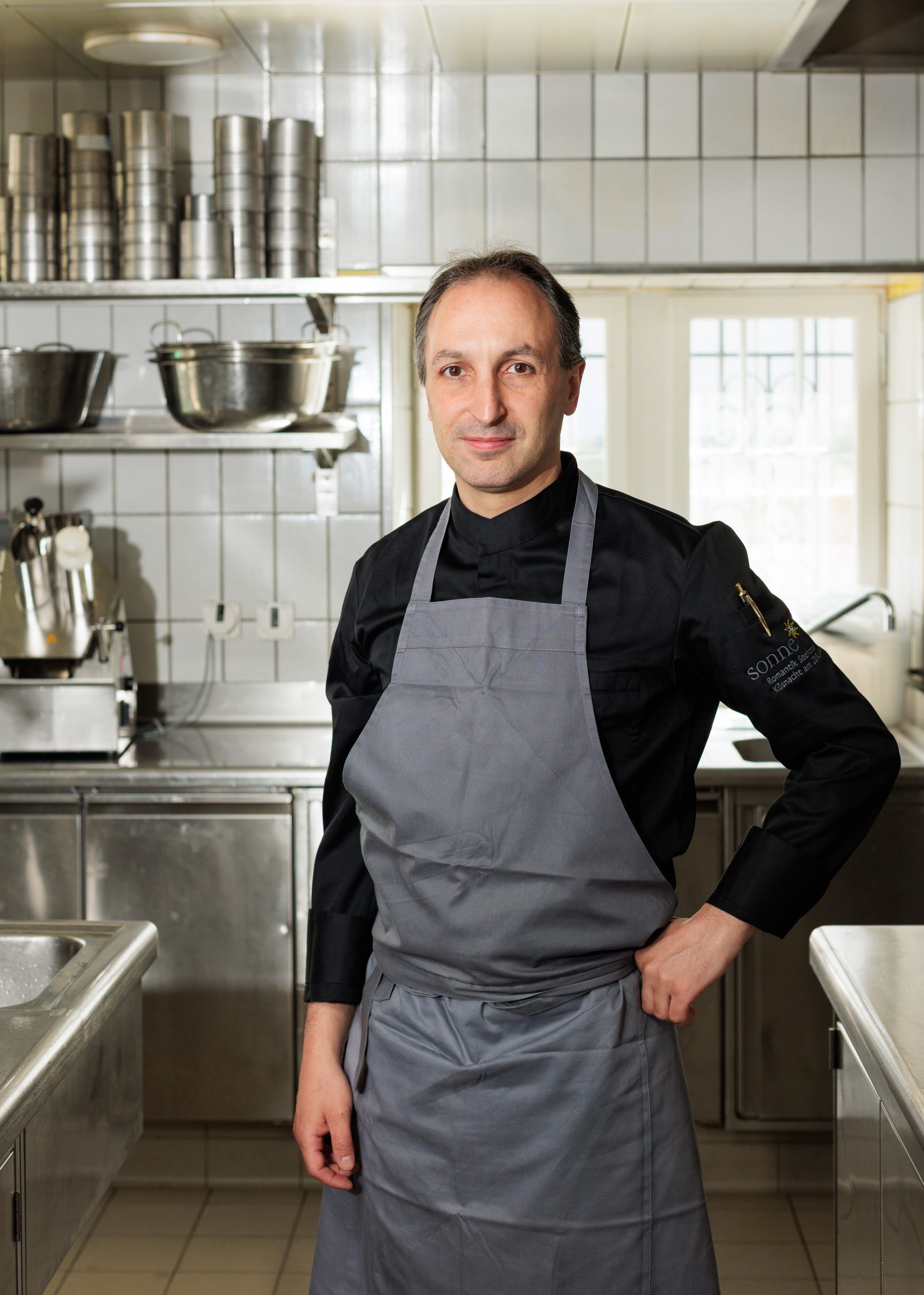 Ein Koch mit schwarzem Hemd und grauer Schürze steht selbstbewusst in einer professionellen Küche mit Edelstahltheken, Regalen und Kochgeräten im Hintergrund.
